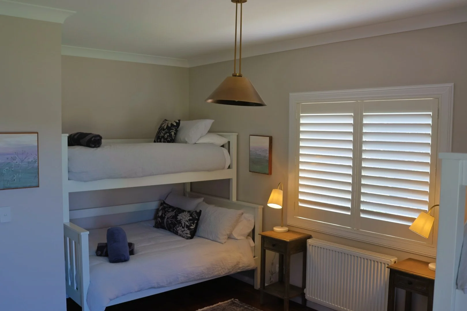Bedroom with white bunk beds, black and white pillows, a small wooden bedside table with a lamp, beige walls, a large window with white shutters, and a ceiling light fixture.