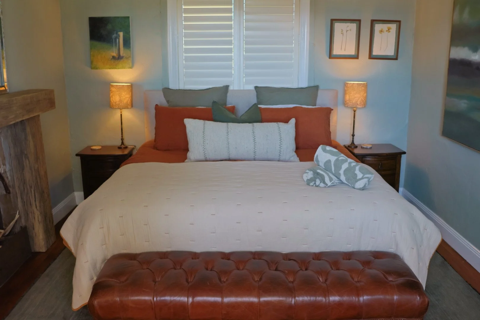 A neatly made bed with a tan quilt, multiple pillows in shades of gray, white, and rust, with a pair of rolled decorative pillows at the foot, flanked by two wooden nightstands with lamps, in a cozy bedroom with paintings and window shutters.
