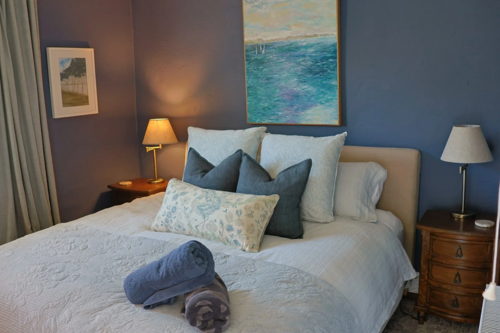 A neatly made bed with a white quilt, several throw pillows in shades of blue and cream, and two rolled towels placed on the foot of the bed. There are two bedside tables with lamps, framed artwork on the walls, and curtains on the window.
