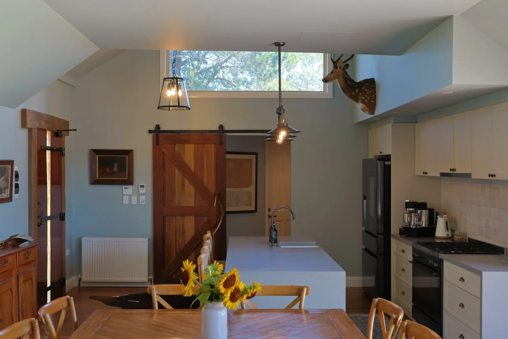 A kitchen with a white island, cream cabinets, and black appliances. A wooden sliding barn door and a vase with sunflowers are on a dining table. There's a deer head mounted on the wall and two pendant lights hanging from the ceiling.