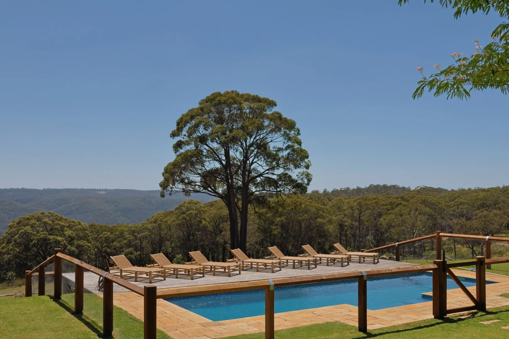 Outdoor swimming pool with wooden lounge chairs, fenced with a railing, on a grassy area with trees and mountains in the background under a blue sky.