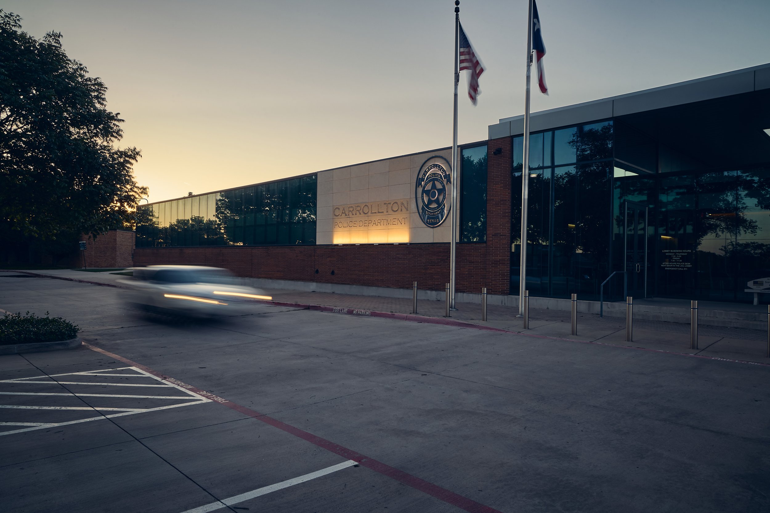 Exterior view of the Carrollton Police Department building at dusk, with flags flying and a blurred car passing by in the parking lot.