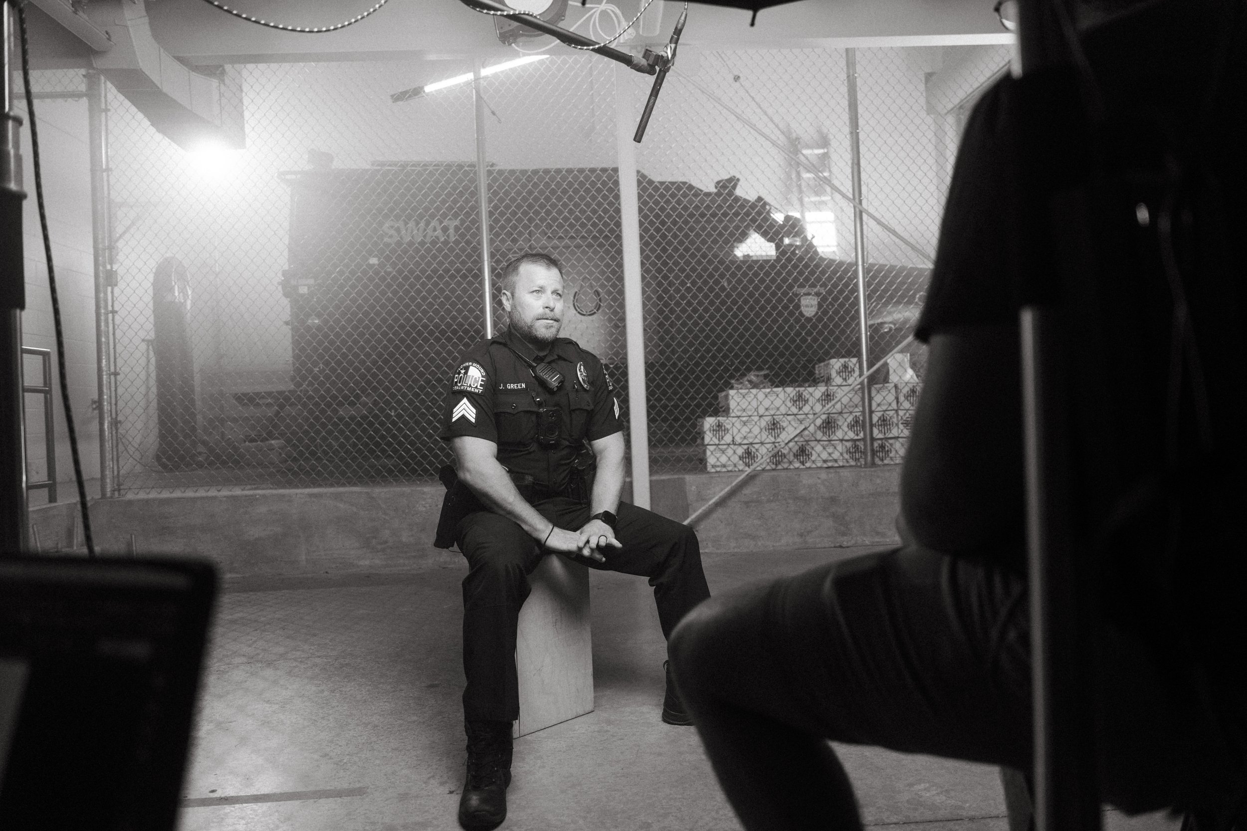Black and white photo of a police officer seated on a stool during an interview or filming, with a chain-link fence and boxes in the background, and another person partially visible in the foreground.