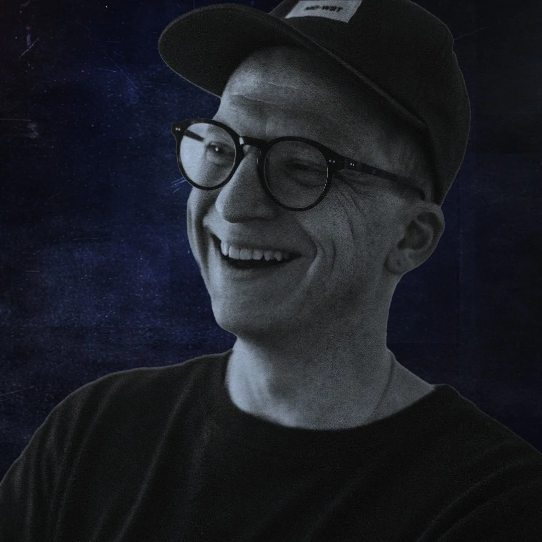A person wearing glasses and a baseball cap, smiling and winking, with a space-themed background.