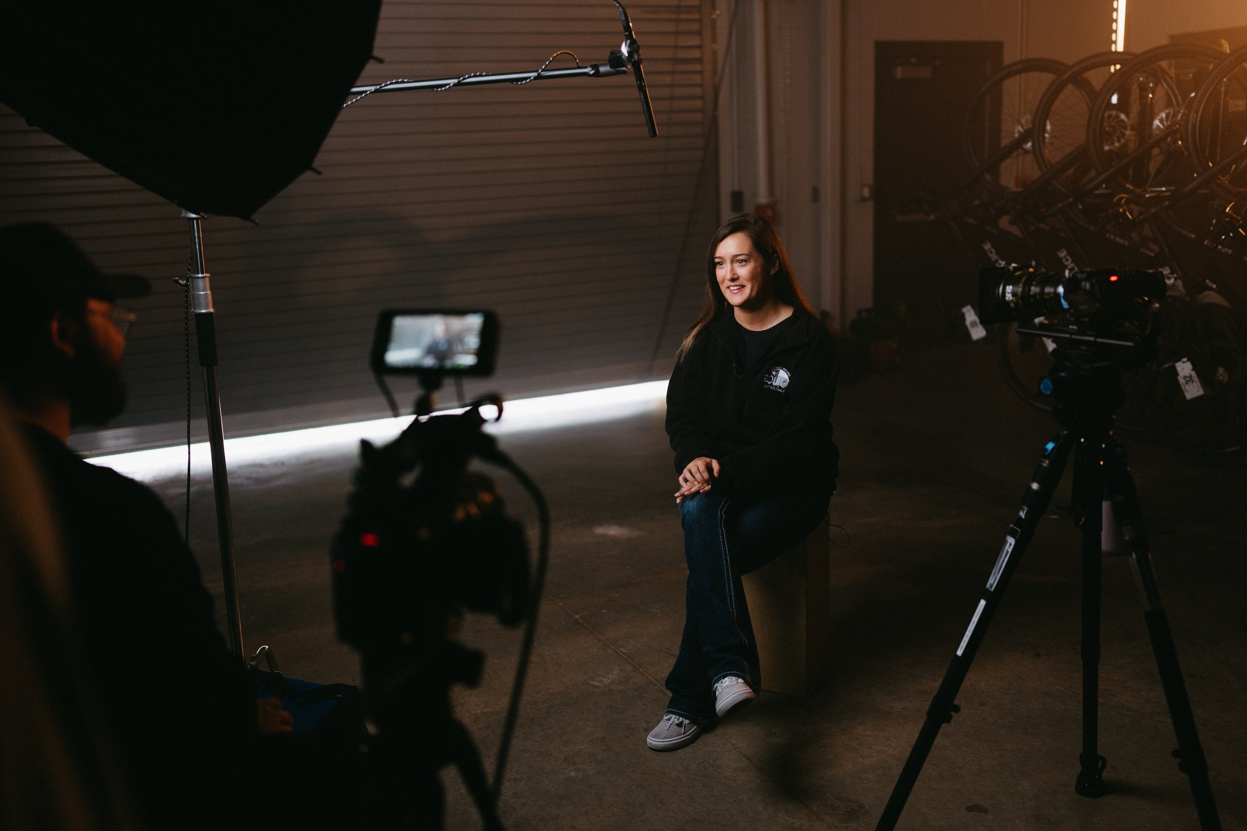 Woman being interviewed in a dimly lit studio with professional camera equipment and a large softbox.