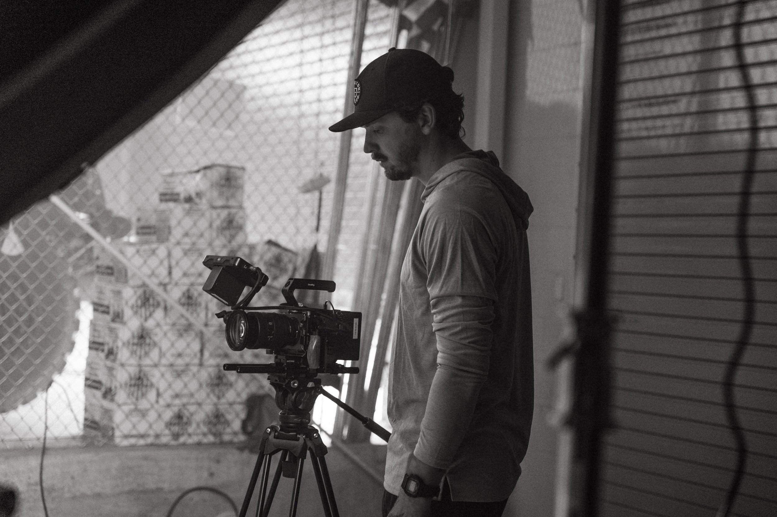 A man with a beard and baseball cap operating a professional movie camera in an outdoor set with chain-link fence in the background.