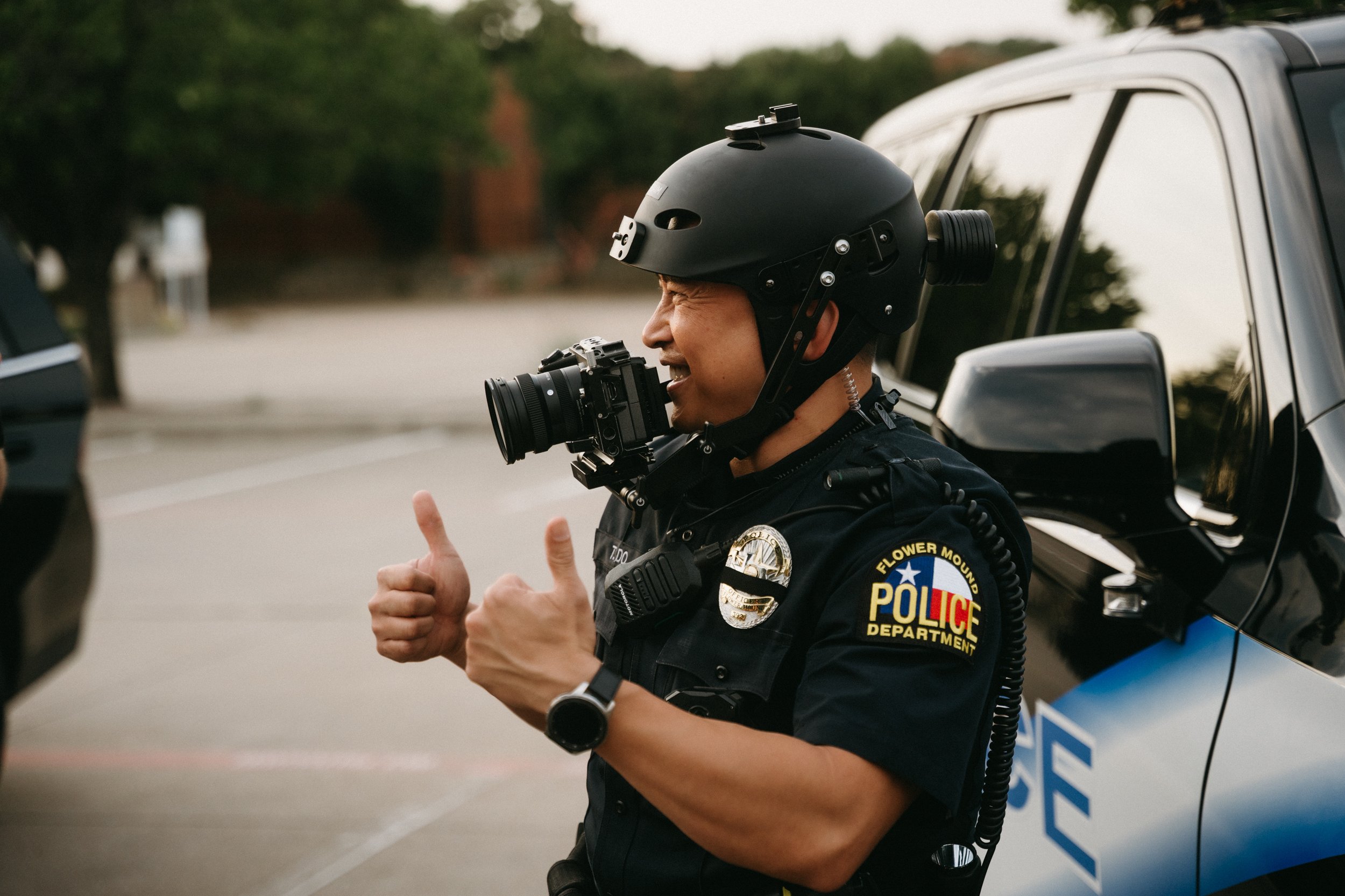 A police officer in tactical gear and a helmet, holding a large camera, giving two thumbs up, standing beside a police vehicle.