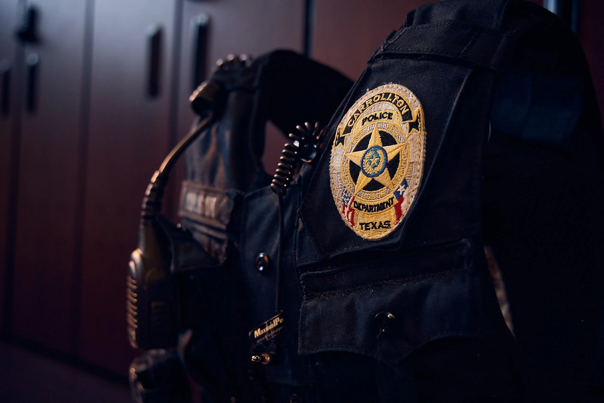 Close-up of a police tactical vest with a badge patch that reads 'Cypress Hill Police Department Texas' hanging on a wall with lockers in the background.