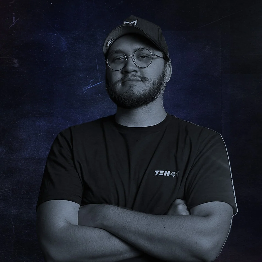 A man with glasses and a beard wearing a black t-shirt and cap, standing with arms crossed against a starry night sky background.