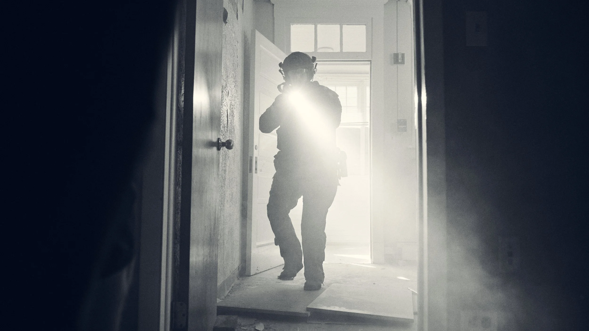 Silhouette of a person in tactical gear, standing in a doorway with bright sunlight behind, creating a backlit effect.