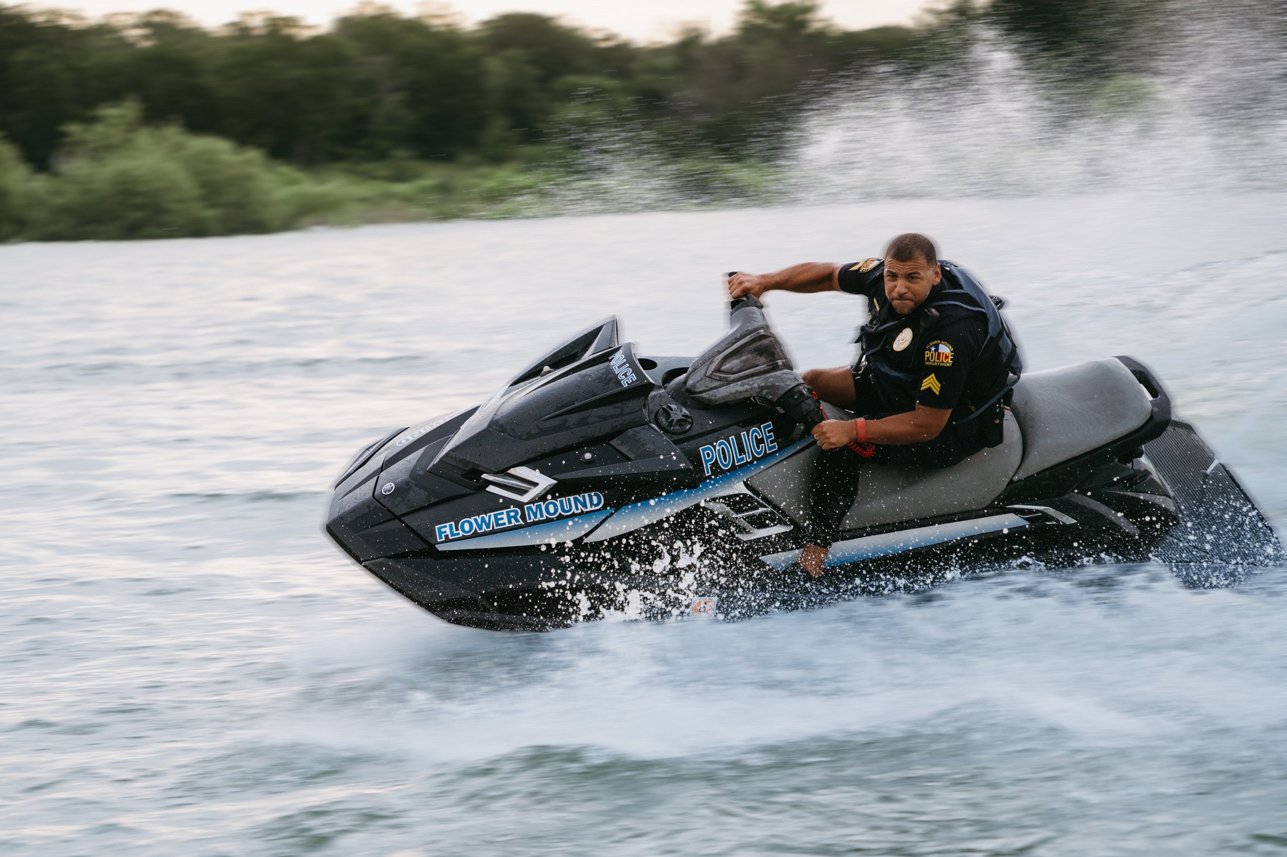 A police officer riding a black jet ski on a body of water, with green trees in the background.