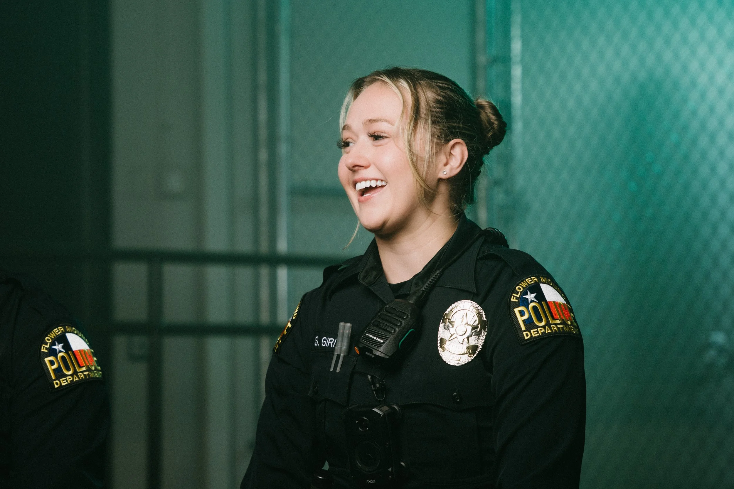 A smiling female police officer with blonde hair in a bun, wearing a black uniform with police patches and a badge, standing in front of a green wall.