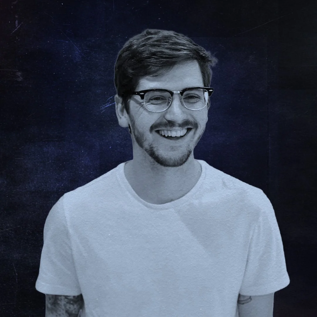 A black-and-white photo of a young man with glasses, facial hair, and a white t-shirt, smiling at the camera against a dark starry background.