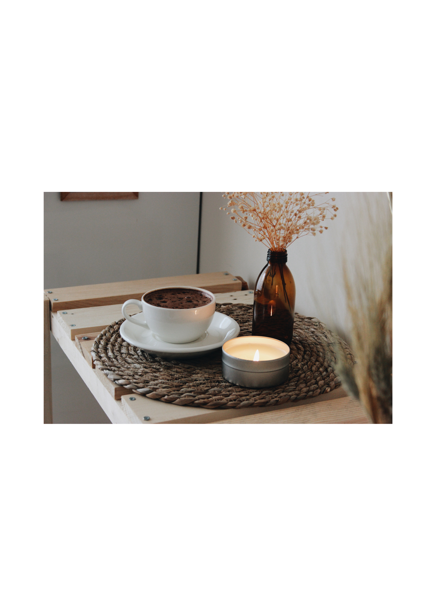 A cup of hot chocolate with a cookie on a saucer, a lit candle, and a dried flowers in a brown glass bottle on a woven placemat on a wooden table.