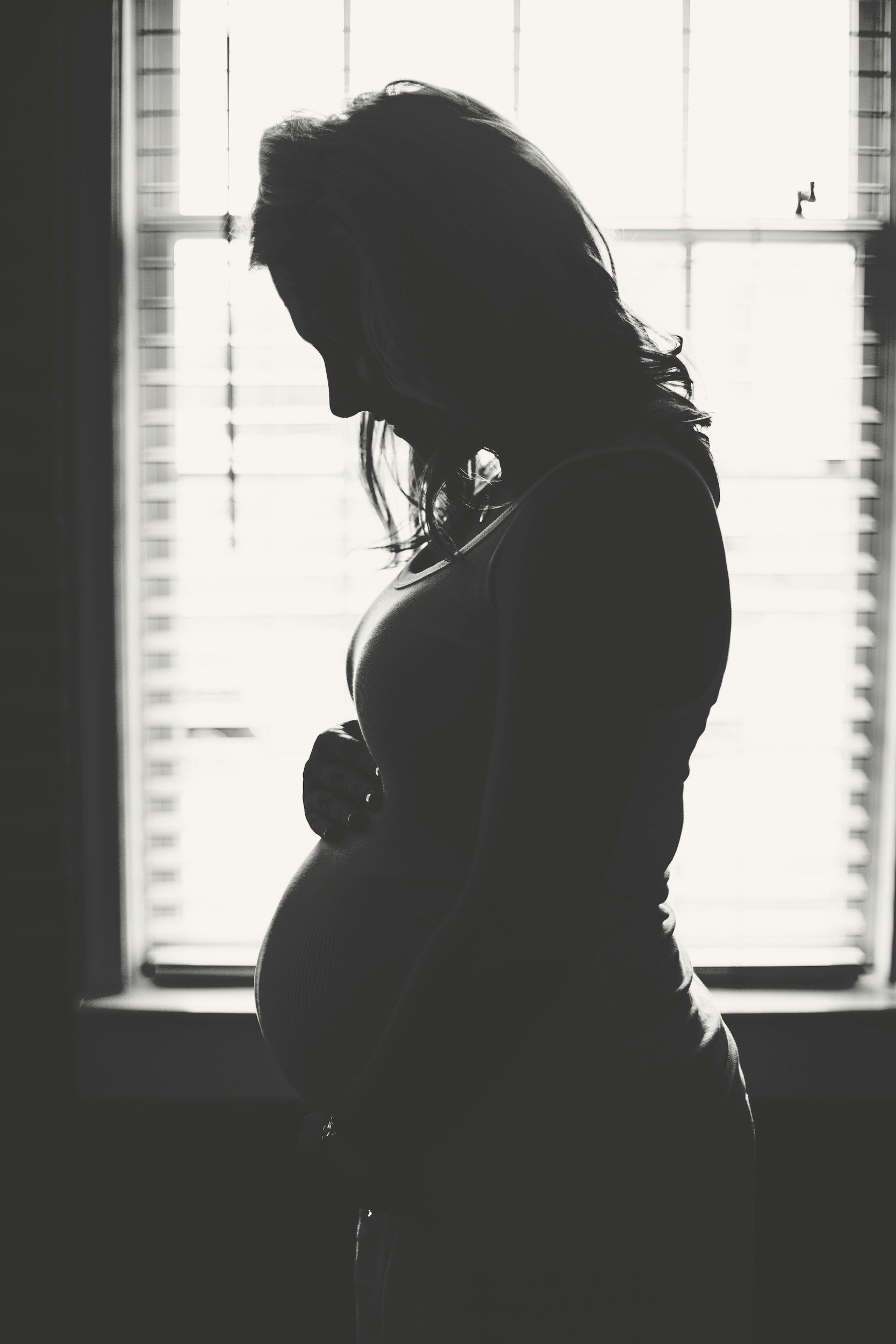 Silhouette of a pregnant woman standing in front of a window with closed blinds, backlit and looking down.
