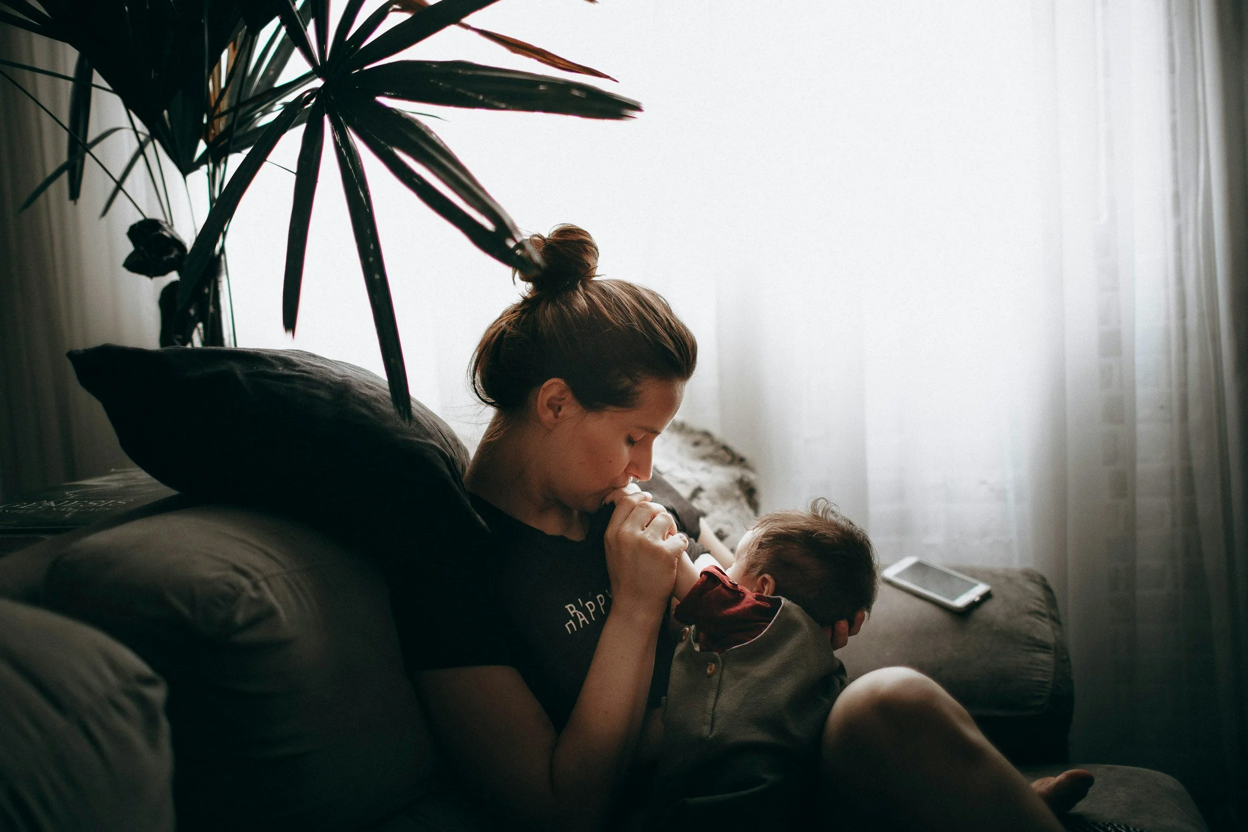 A woman sitting on a couch in a dimly lit room, drinking from a cup while holding a young child. There is a large plant nearby and a smartphone on the couch next to her, with sheer curtains and bright light coming through the window in the background.