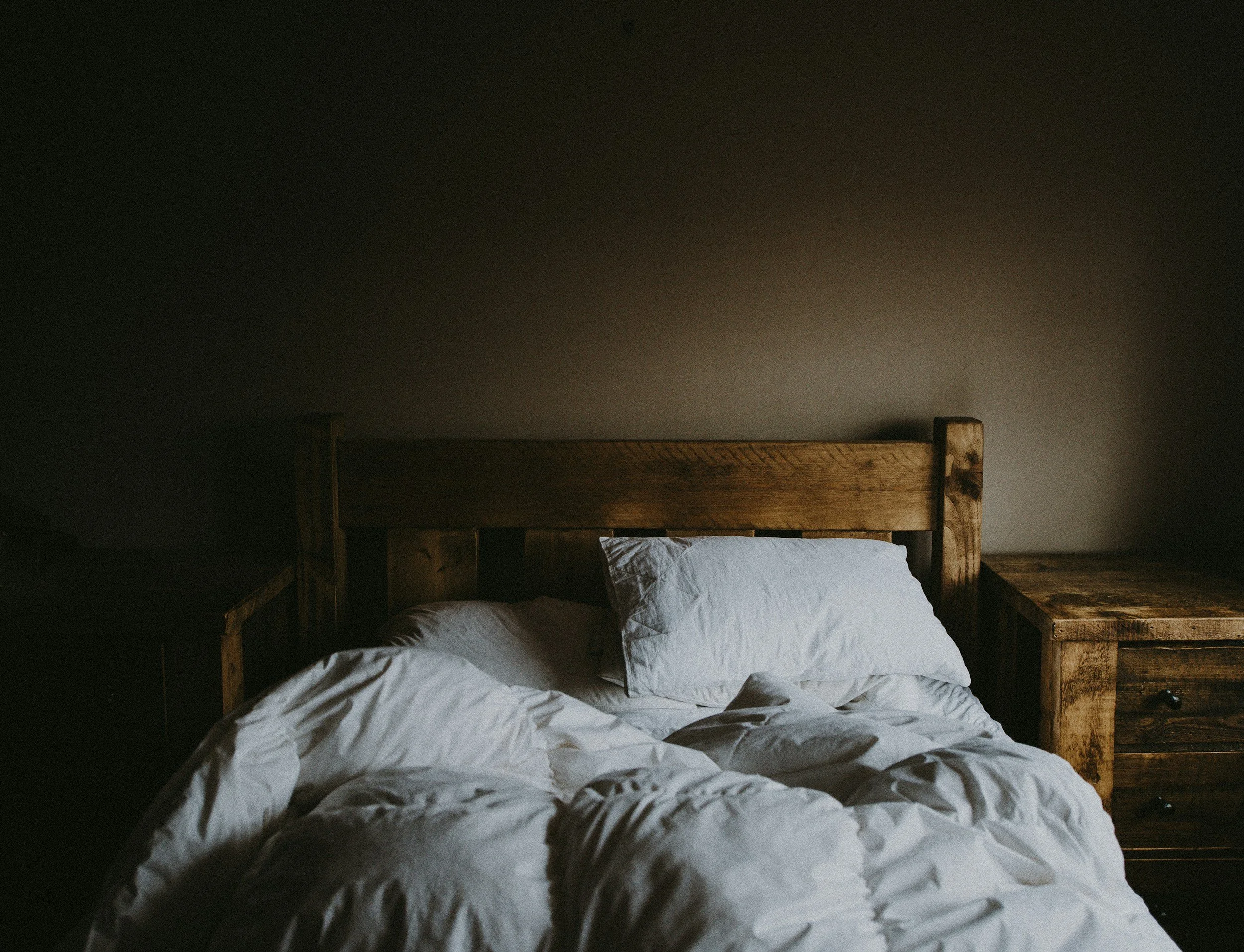 Unmade bed with white sheets and pillows, wooden headboard, and nightstand, in a dimly lit bedroom.