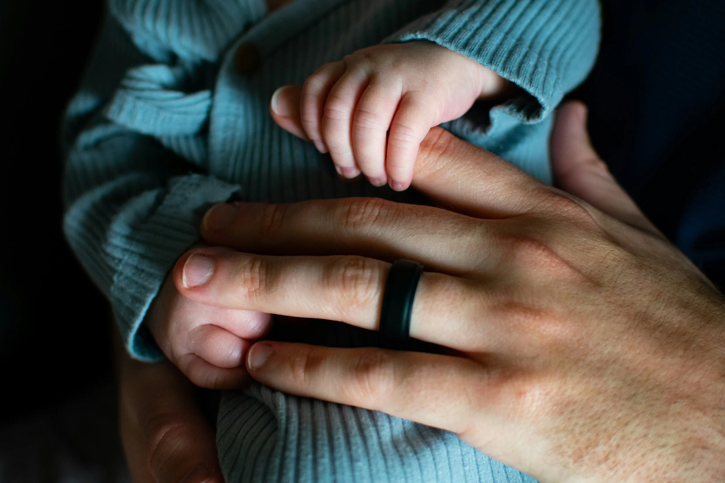 Close-up of an adult's hand holding a baby's hand, both with their fingers intertwined, wearing a ring on the adult's finger.