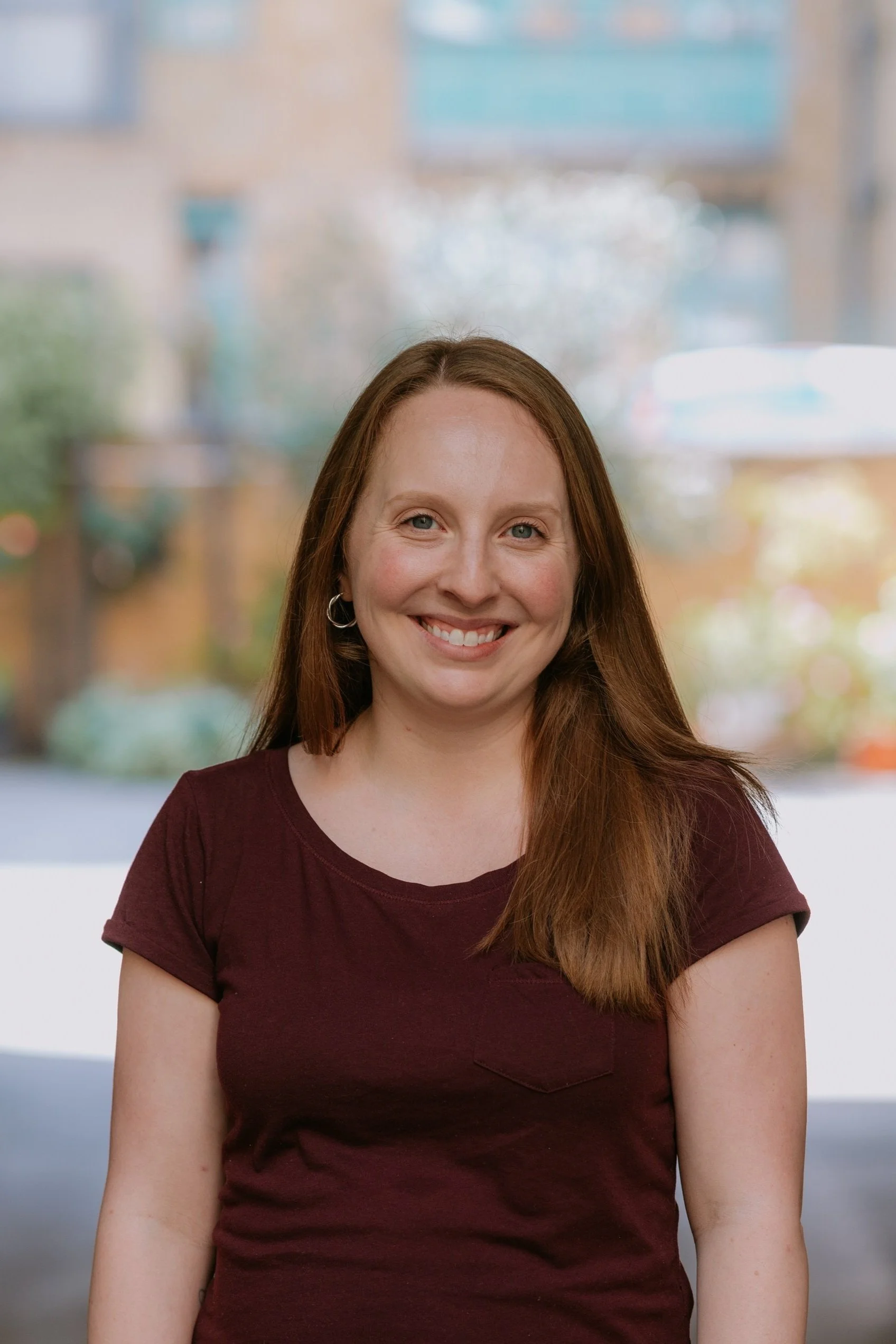 A smiling woman with long red hair wearing a maroon t-shirt standing outdoors with blurred trees and buildings in the background.
