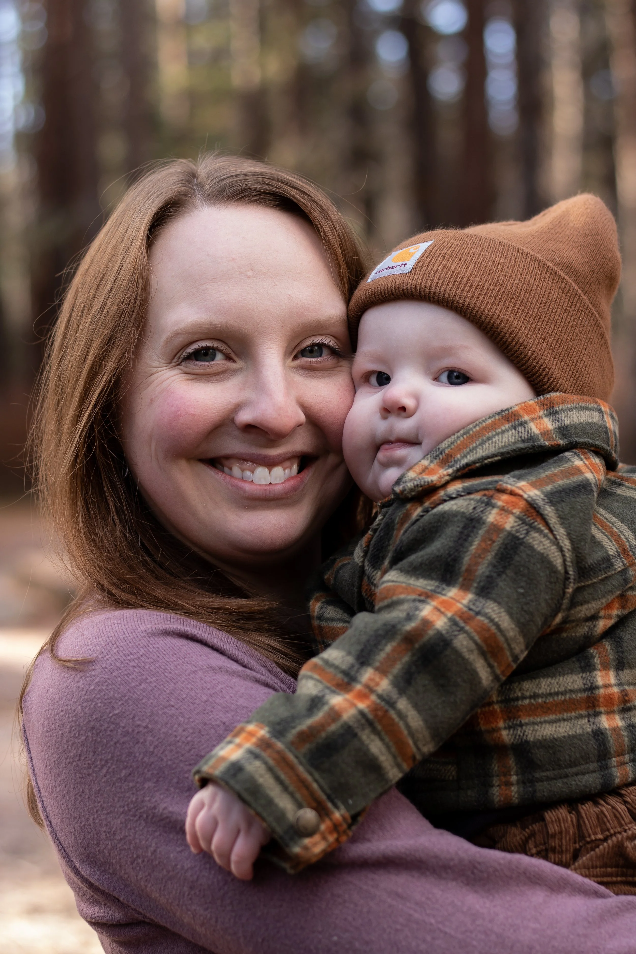 A woman with red hair smiling while holding a baby in a forest setting during daylight. The baby is wearing a brown beanie and a plaid coat, looking at the camera.