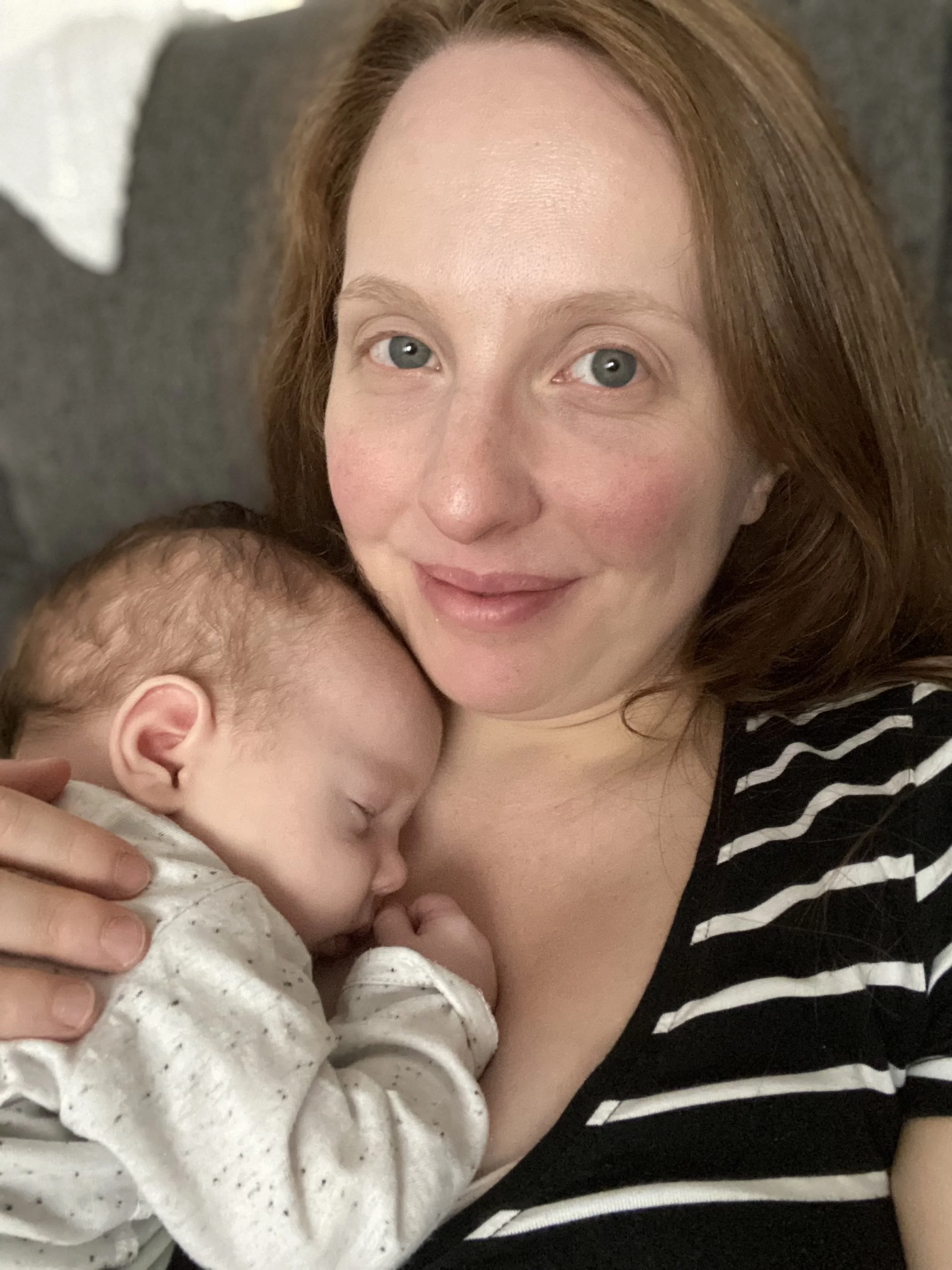 A woman with red hair and a striped shirt holding a sleeping baby with red hair, both cozy and content.