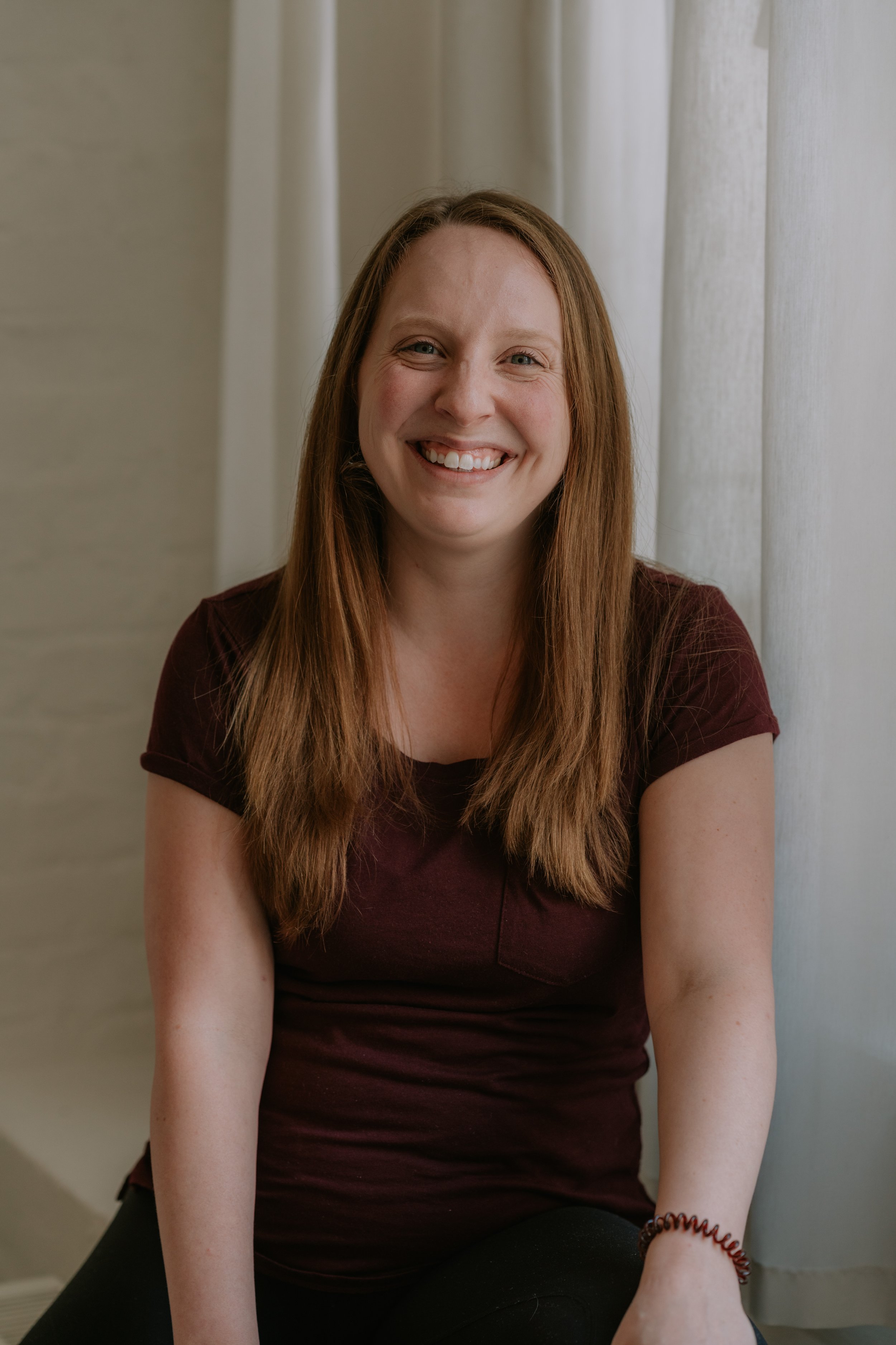 A smiling woman with long red hair sitting in front of white curtains.