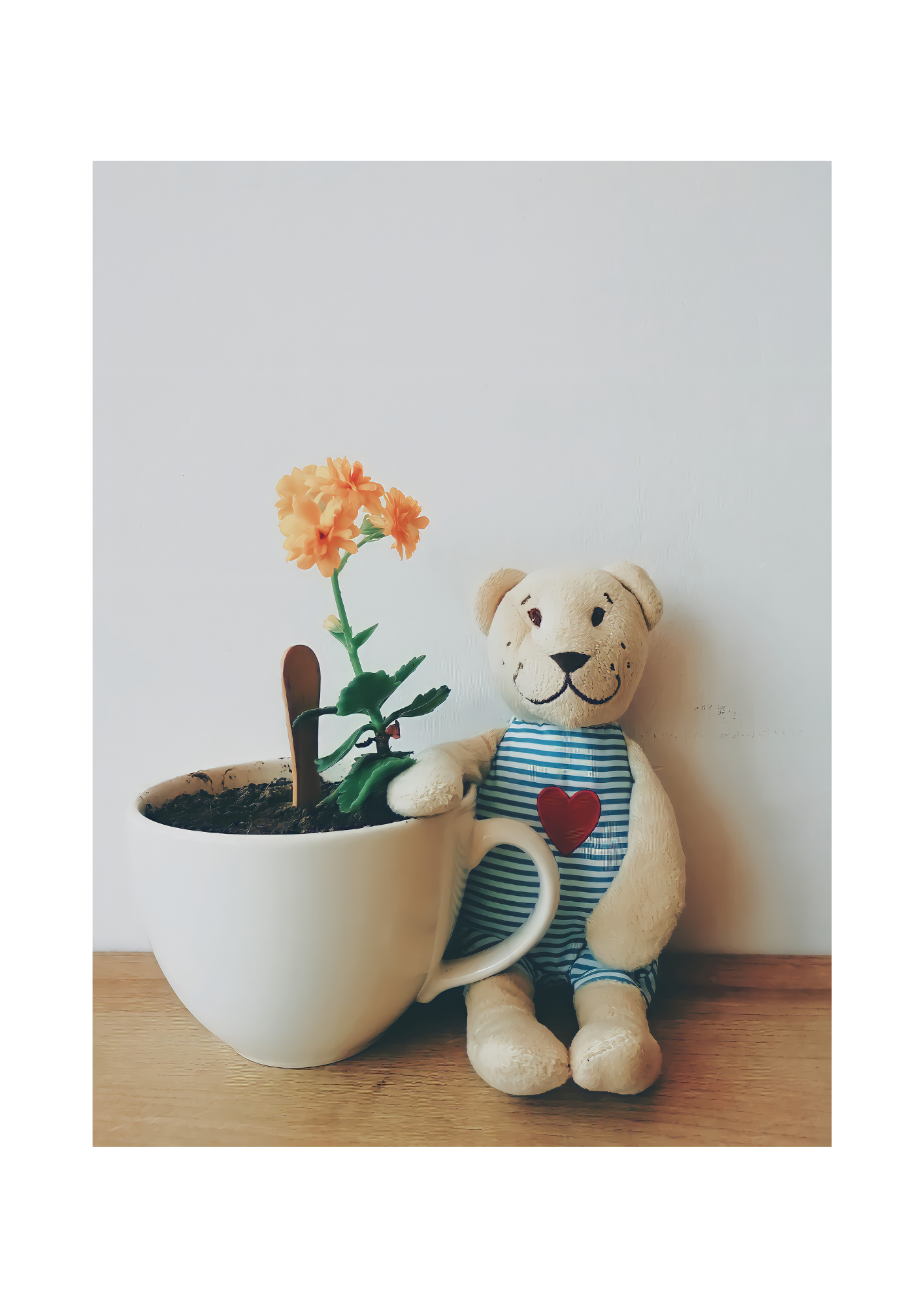 A plush teddy bear wearing a striped shirt with a red heart, sitting next to a white teacup-shaped planter with an orange artificial flower, on a wooden surface against a white wall.