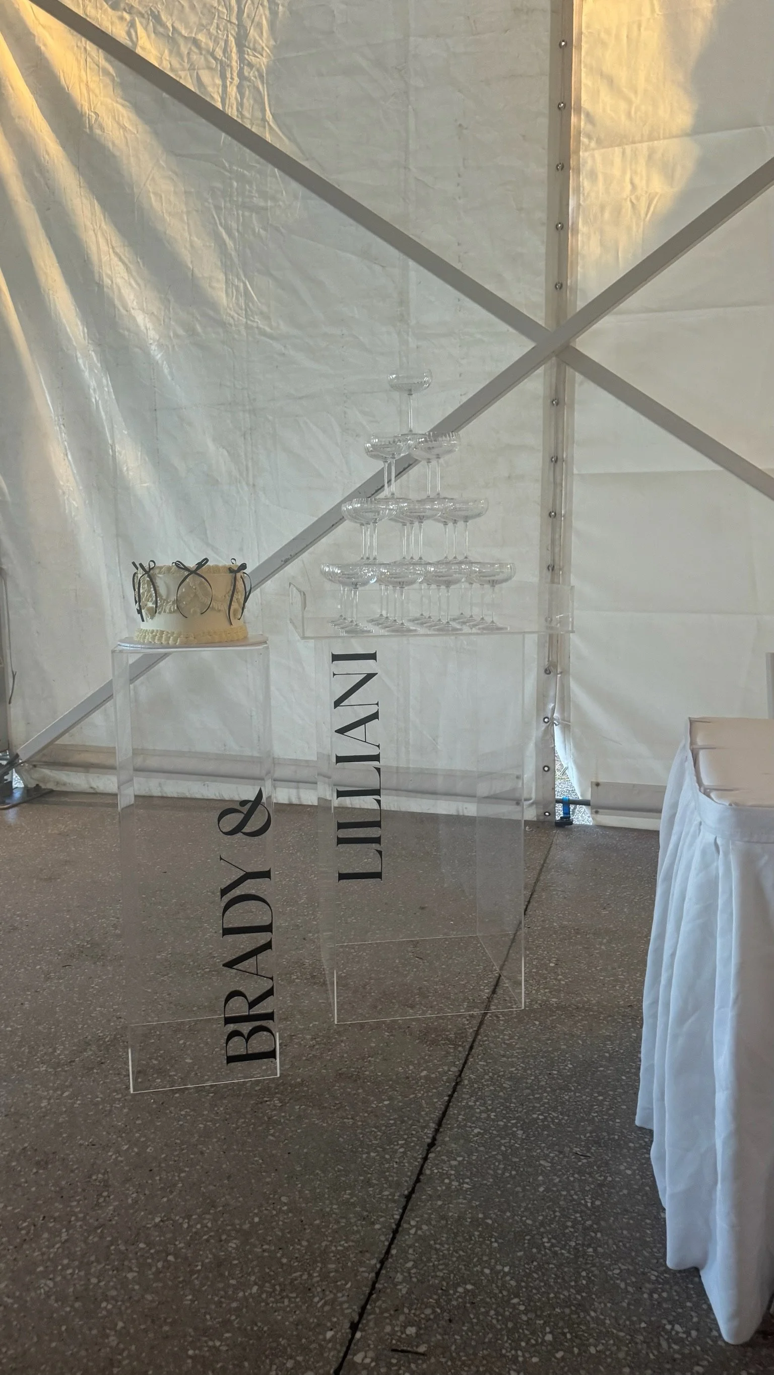 A wedding cake and a champagne tower on a clear acrylic table with the words 'BRIDE & BRIDESMAID' printed on it, set inside a white tent.