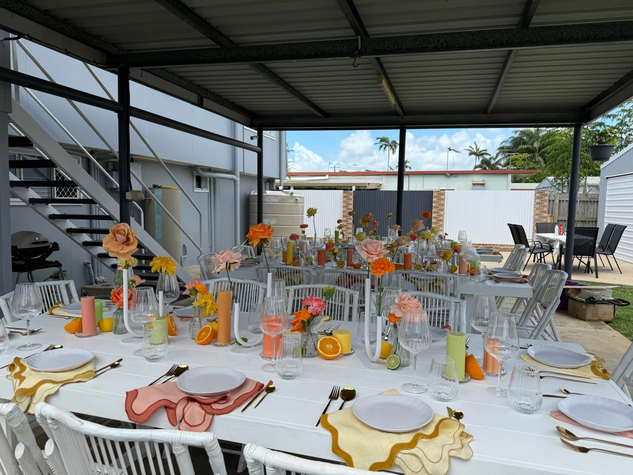 Outdoor event setup with long tables decorated with pink, orange, and yellow flowers, fruit slices, candles, wine glasses, and gold cutlery, under a metal-roofed shelter.