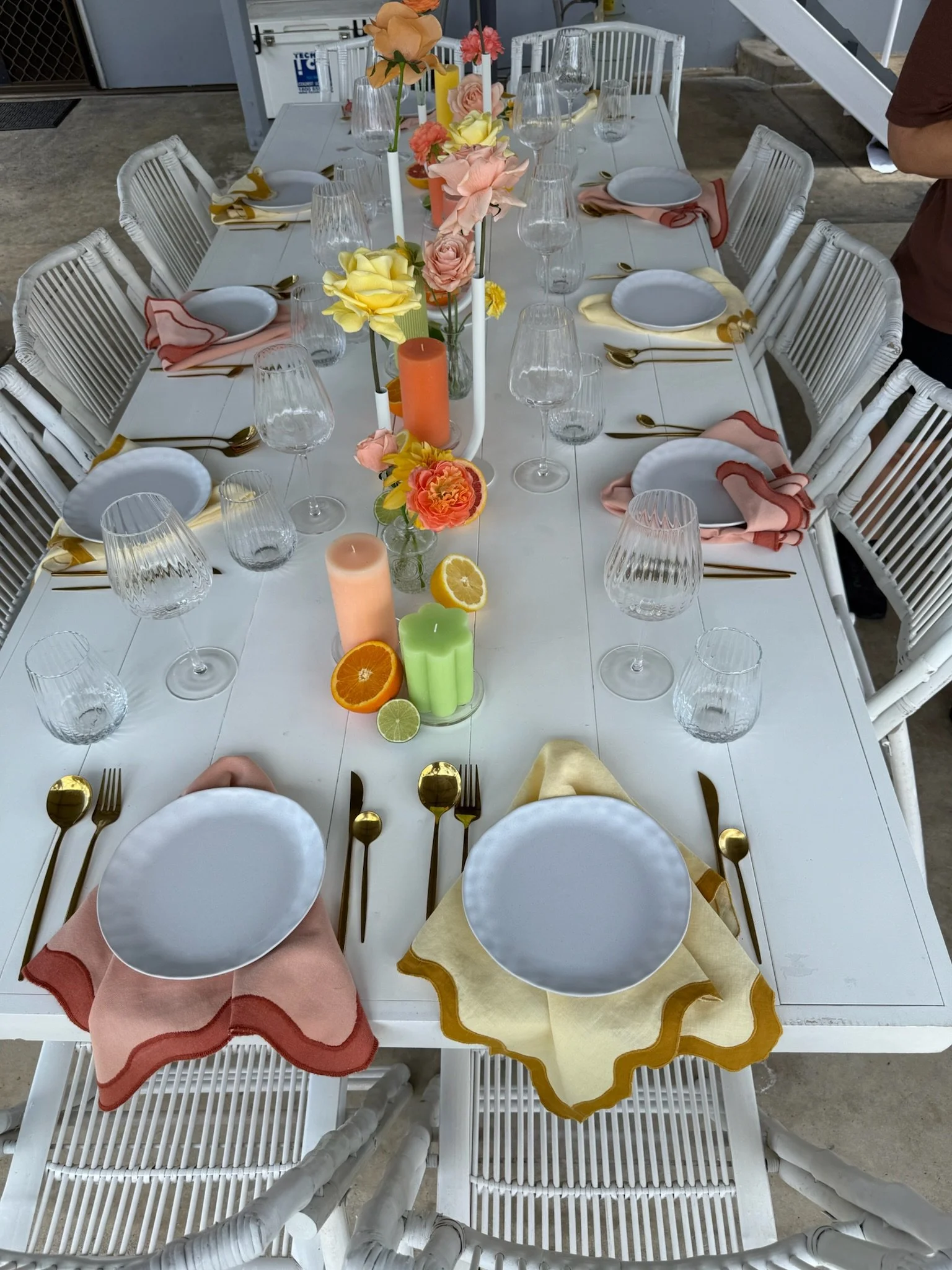 A rectangular dining table set for a meal with white plates, gold flatware, pink and yellow napkins, and clear wine glasses. The table has a floral centerpiece with pastel-colored flowers and decorative candles in various colors. The setting suggests