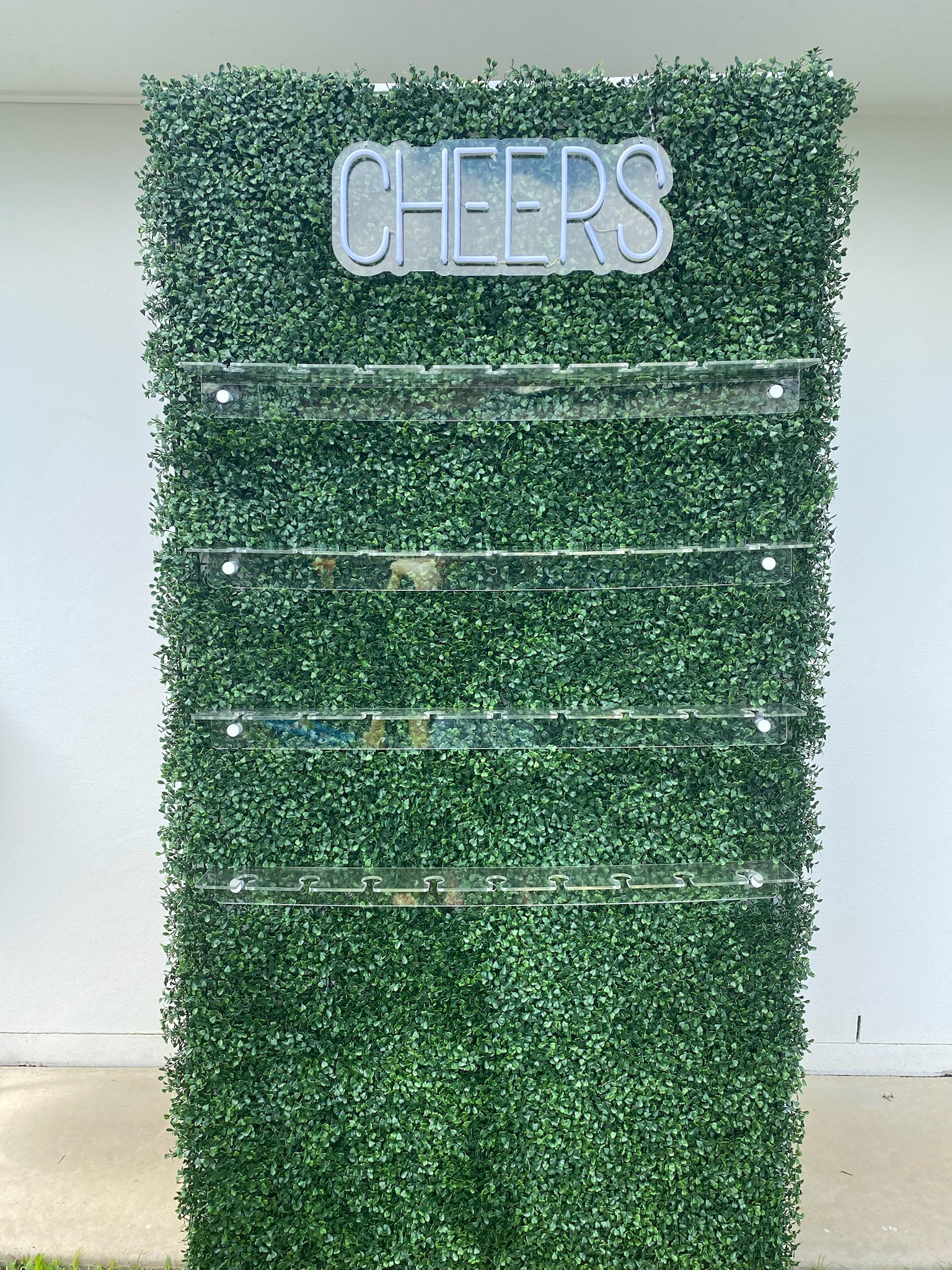 Green hedge wall with a neon sign that says 'CHEERS' at the top and empty clear shelves below.