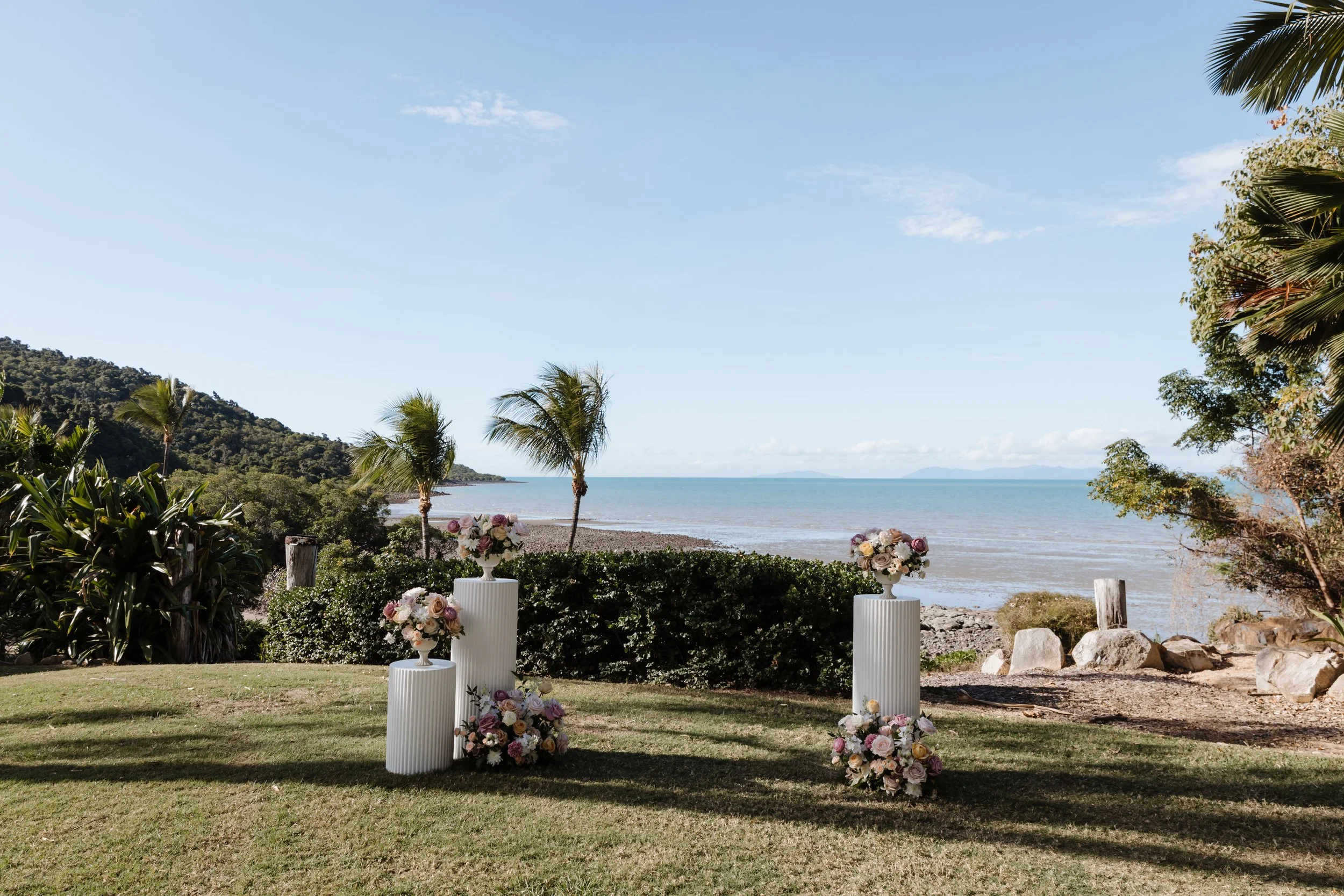 Beachside wedding setup with three white cylindrical pedestals topped with floral arrangements, overlooking the ocean with palm trees and lush greenery in the background.