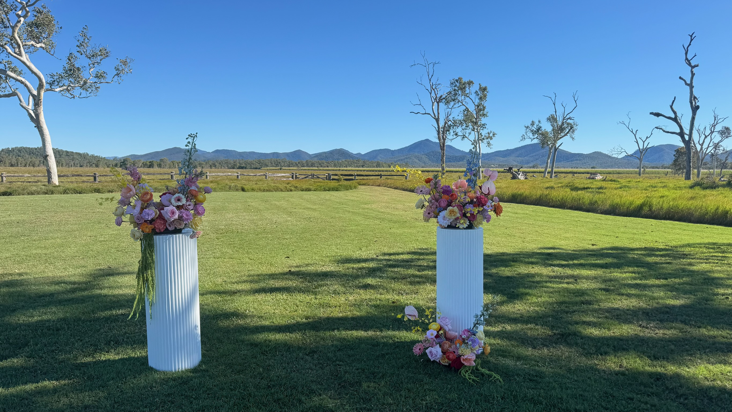 Two tall white vases filled with pink, purple, white, and yellow flowers stand on a green lawn, with a scenic background of trees, mountains, and a blue sky.