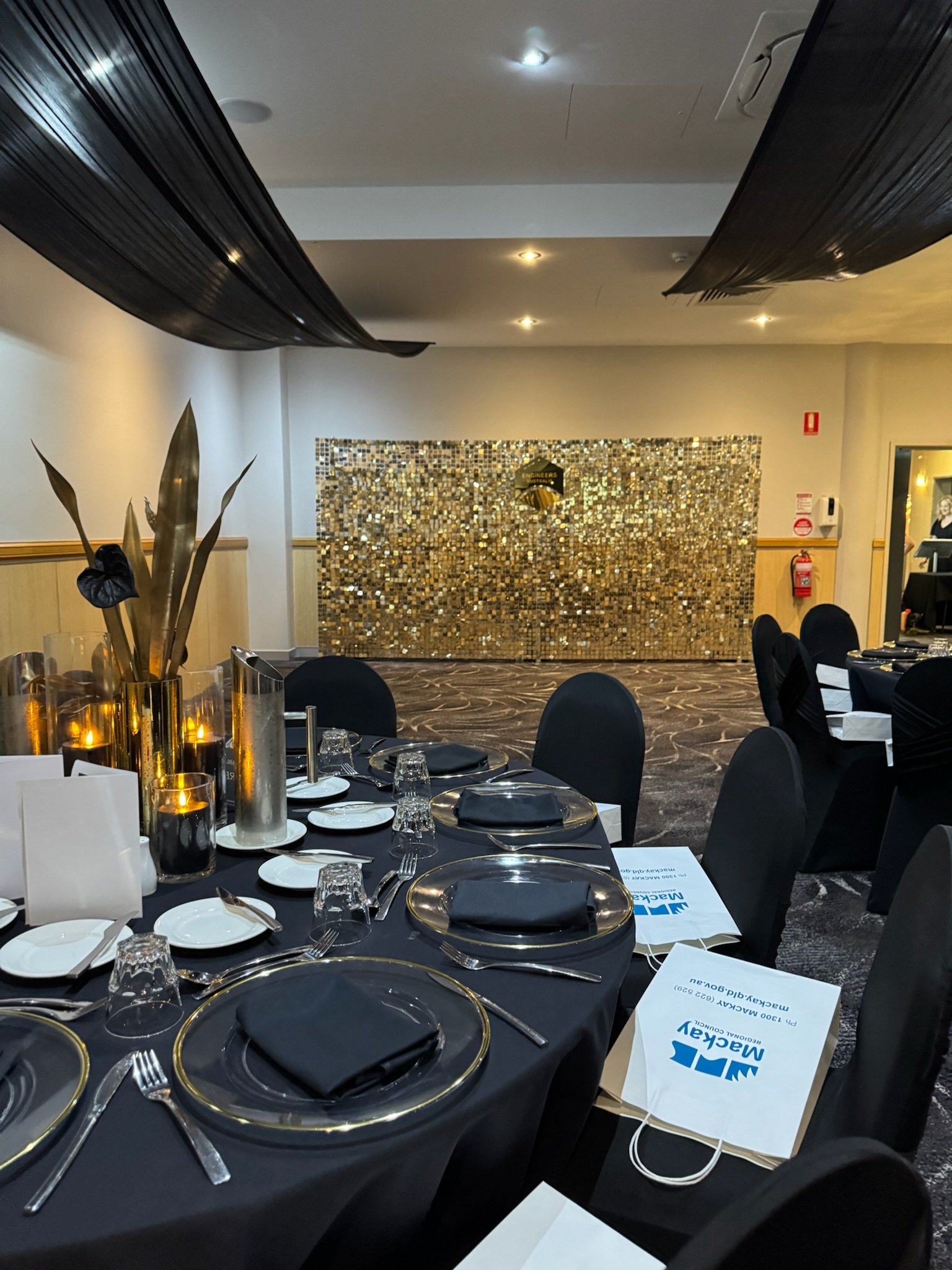 Round banquet table set for a formal event with black chairs, black napkins, gold-rimmed plates, and glassware, decorated with candles and gold accents, with a gold mosaic backdrop in the background.
