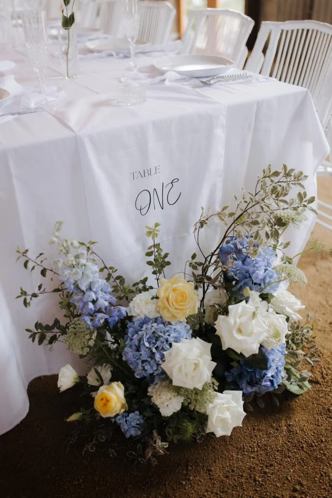 A table decorated with a white tablecloth, table settings, and a floral arrangement at a wedding or event. The table sign indicates 'Table One'.