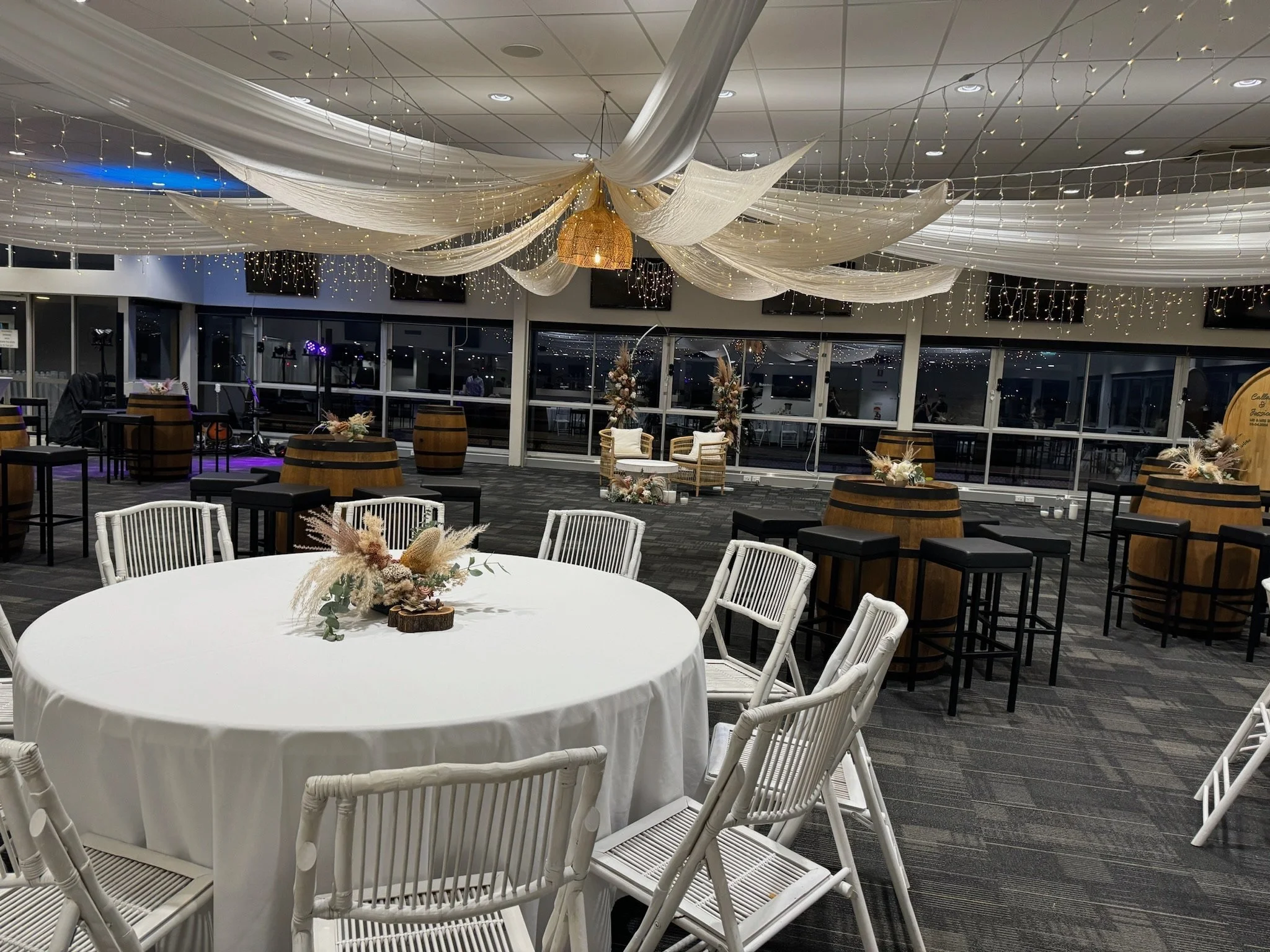 Decorated event space with a round table, white chairs, hanging fabric, string lights, and barrel tables with floral centerpieces.
