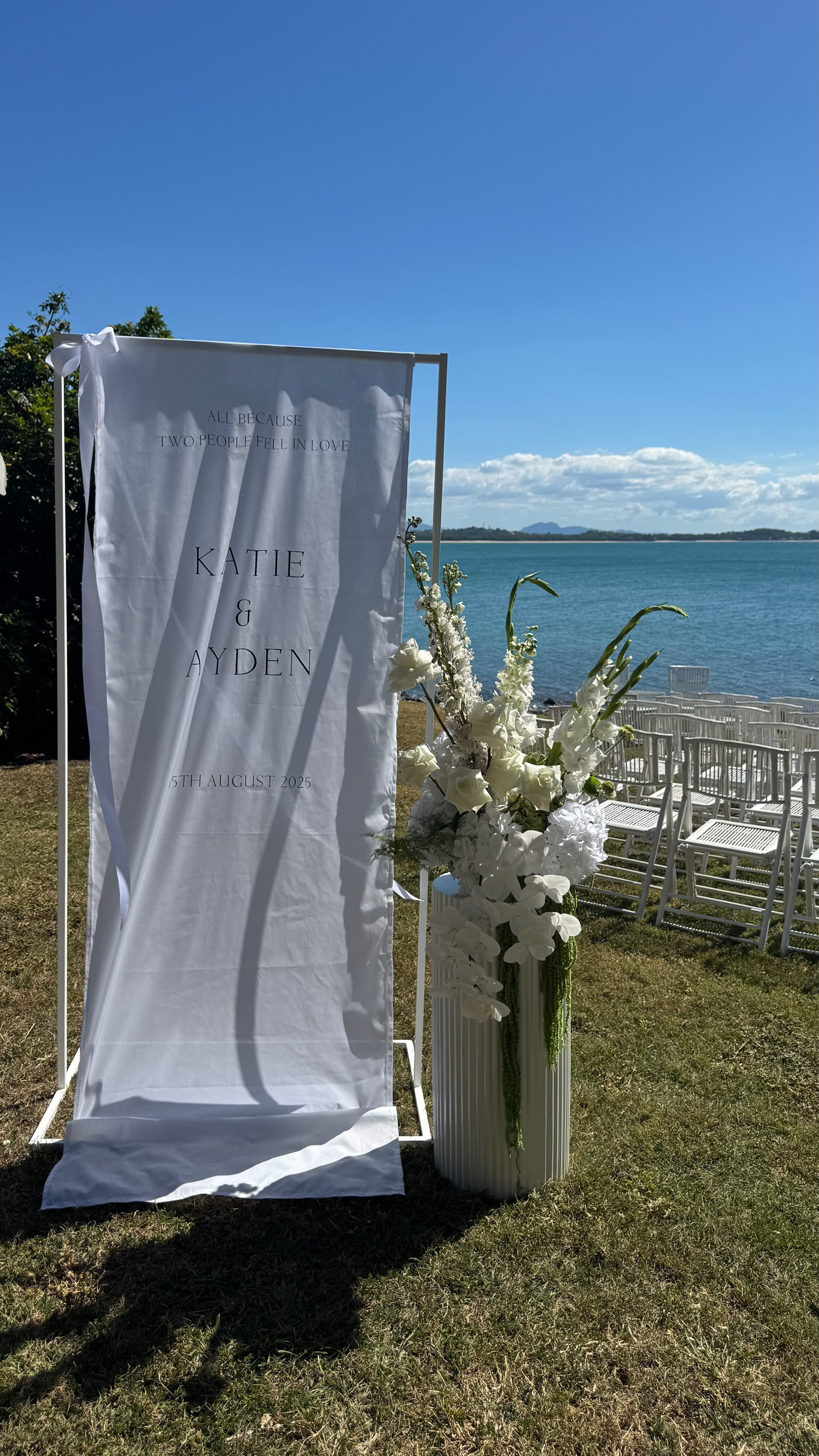 Wedding setup near a body of water with a white floral arrangement and a fabric banner with text, sunny sky and chairs in the background.