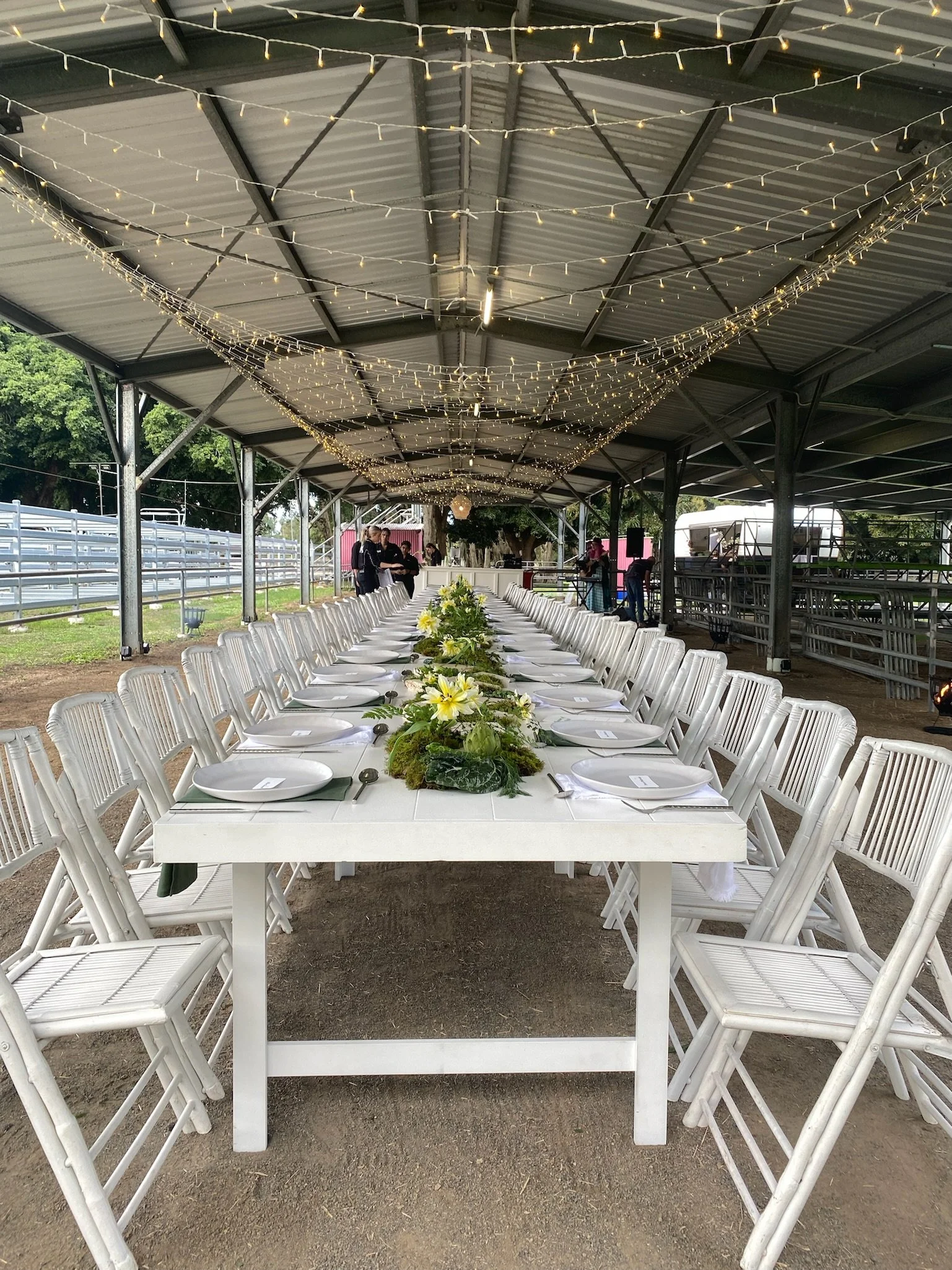 Long rectangular table set for an outdoor event under a metal shelter, decorated with floral arrangements and surrounded by white chairs, with string lights hanging from the ceiling.