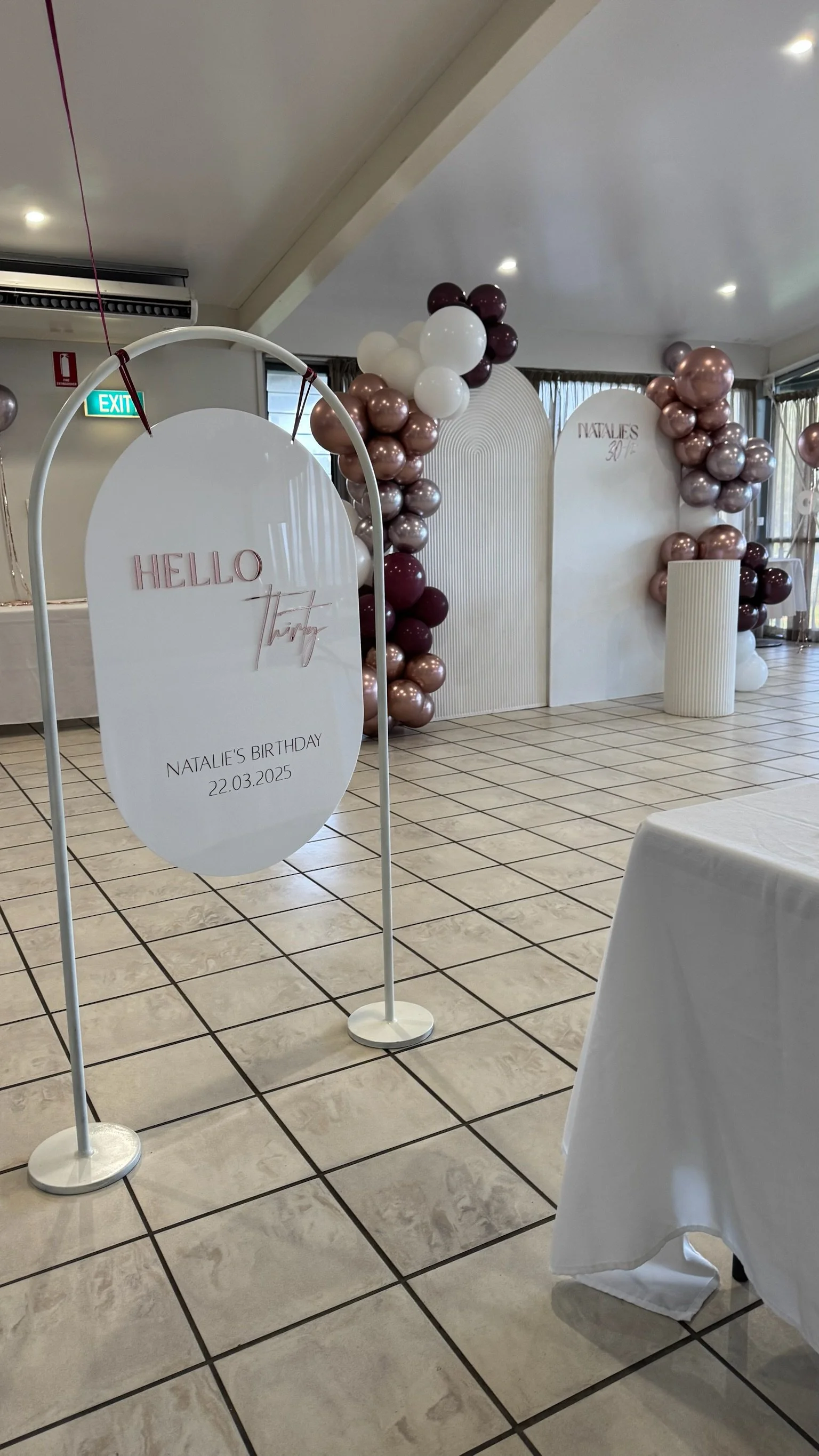 Decorated birthday party area with balloon arrangements in shades of white, pink, and purple, featuring a white arch and panels with text announcing Natalie’s birthday on March 22, 2025.