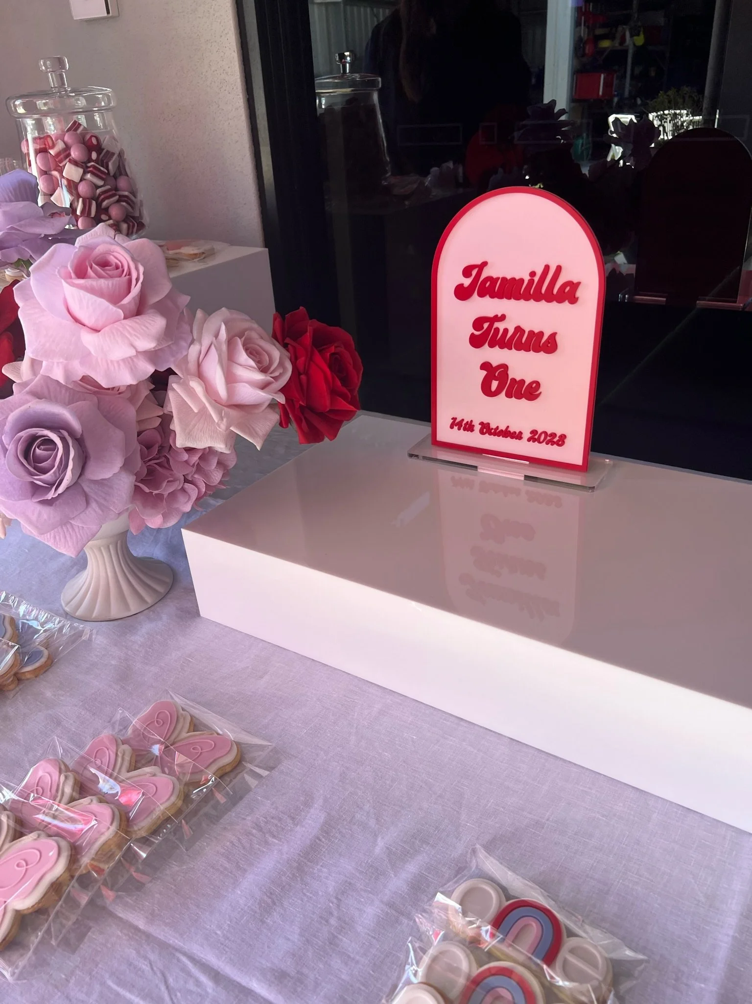 A table with pink and purple paper roses in a white vase, decorated cookies in clear packaging, and a pink sign that reads 'Jamilya Turns One 14th October 2023'.