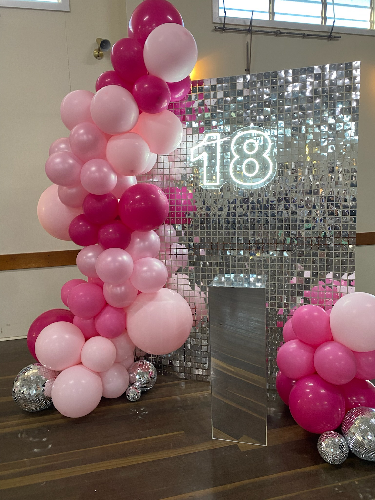 Decorative setup with pink and magenta balloons, silver disco balls, a mirrored tile backdrop, and a neon sign displaying the number 18 for an 18th birthday celebration.