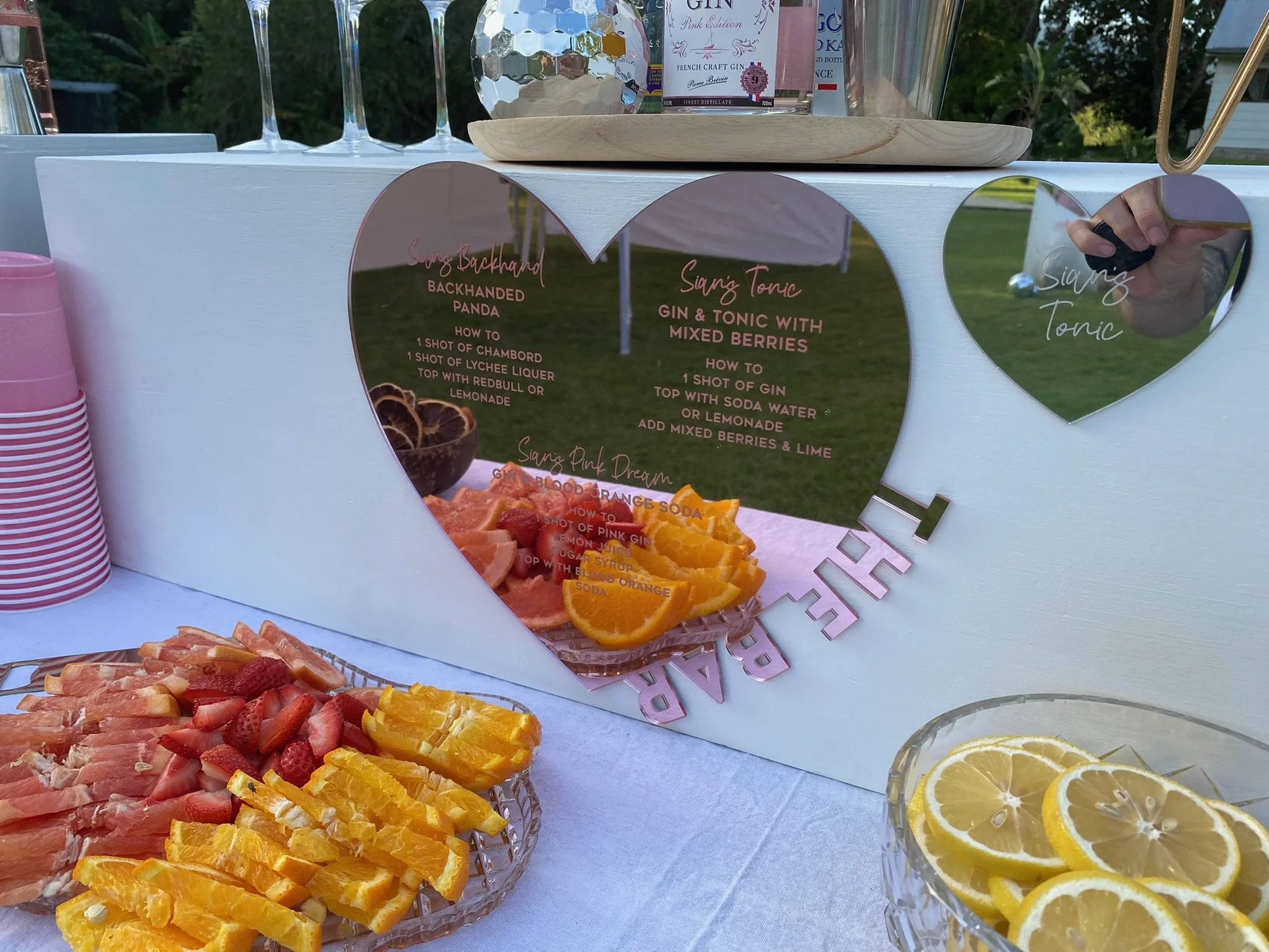 A table at an outdoor event with slices of oranges, strawberries, and grapefruit on glass platters, a reflective heart-shaped sign with recipes for signature drinks, and pink paper cups stacked on the side.