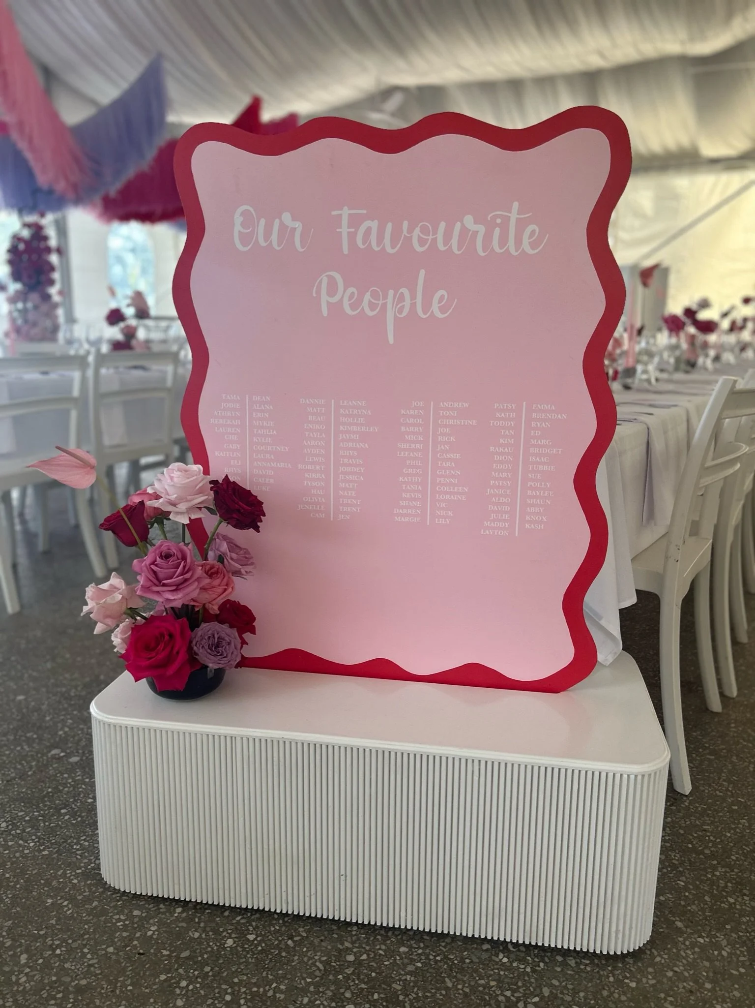 A pink welcome sign with the title 'Our Favourite People' and a list of names underneath, set on a white stand, decorated with a bouquet of pink and red roses.