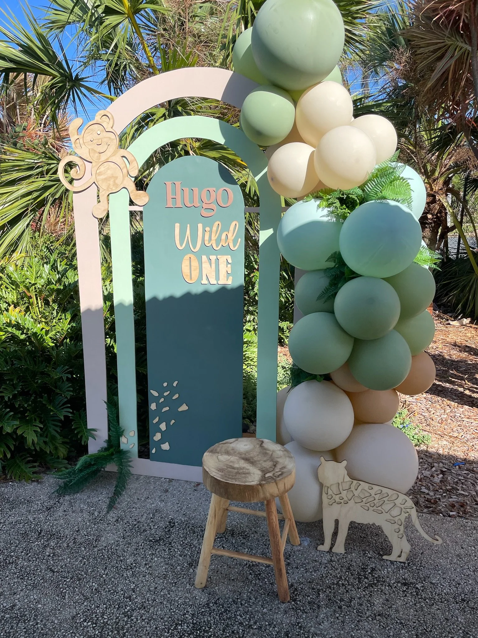 Decorative photo booth setup for a first birthday celebration with a pastel arch, balloons in shades of green, blue, and cream, a wooden stool, a cutout of a lion, and a sign that reads 'Hugo Wild ONE' surrounded by greenery.