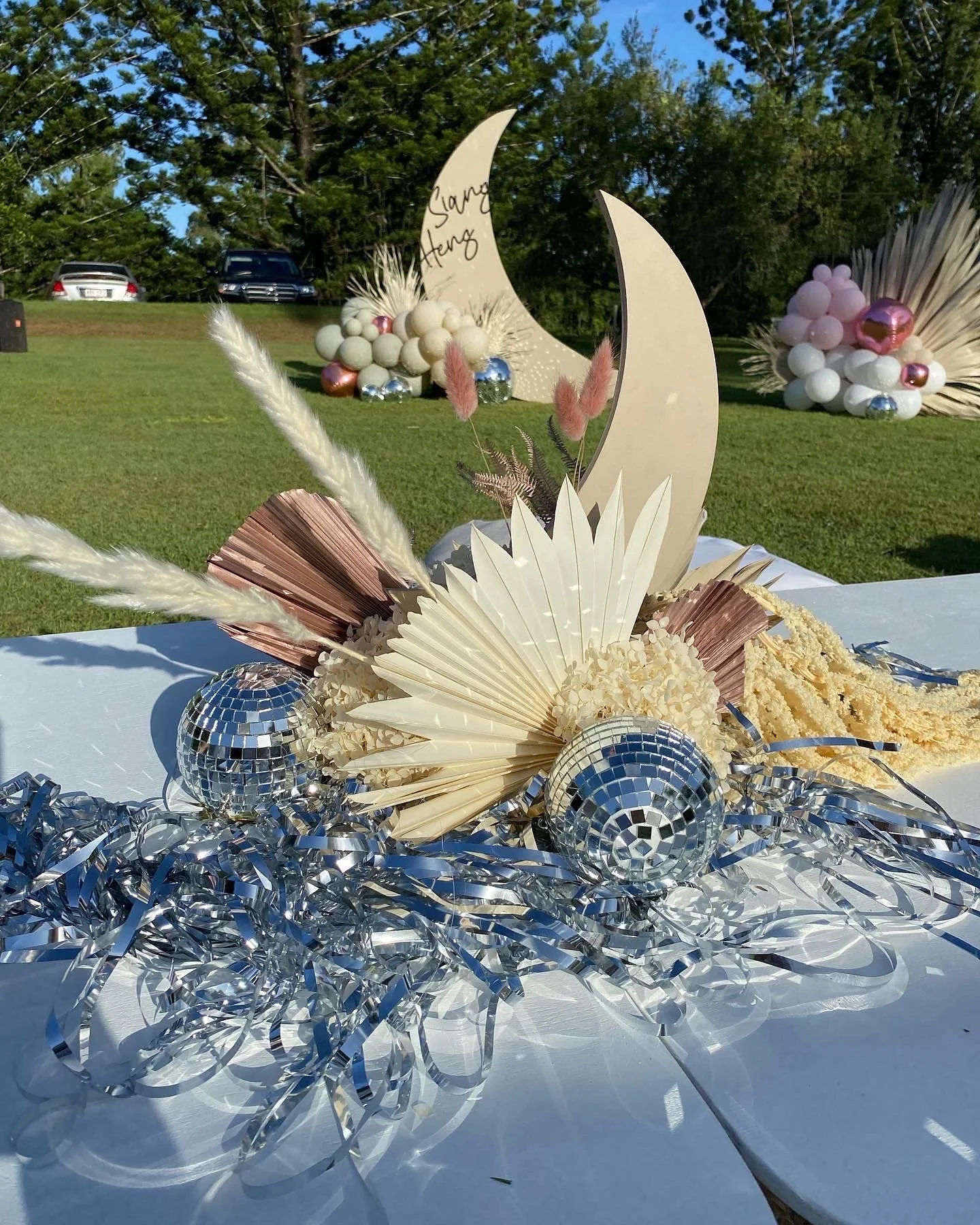 Decorative outdoor table centerpiece with white and pink dried flowers, crescent moon and star shapes, small disco balls, and metallic silver streamers, set against a grassy background with balloons and trees.