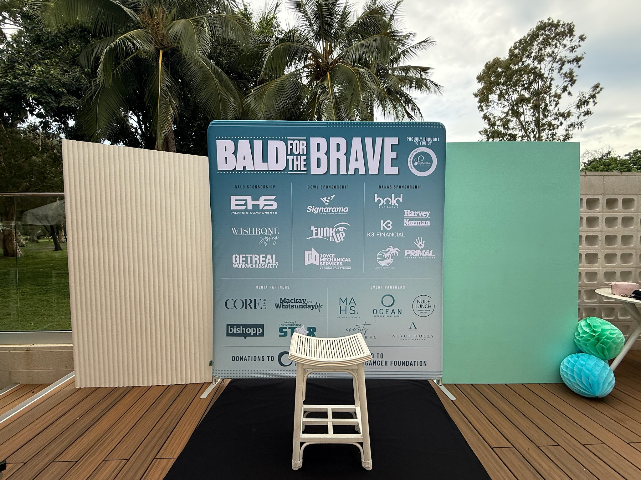 Event display for 'Bald for the Brave' fundraiser featuring sponsor logos on a large blue board, with paper lanterns in green and blue on the right, a white chair in front, and decorative panels on each side, set outdoors on a wooden deck with trees 