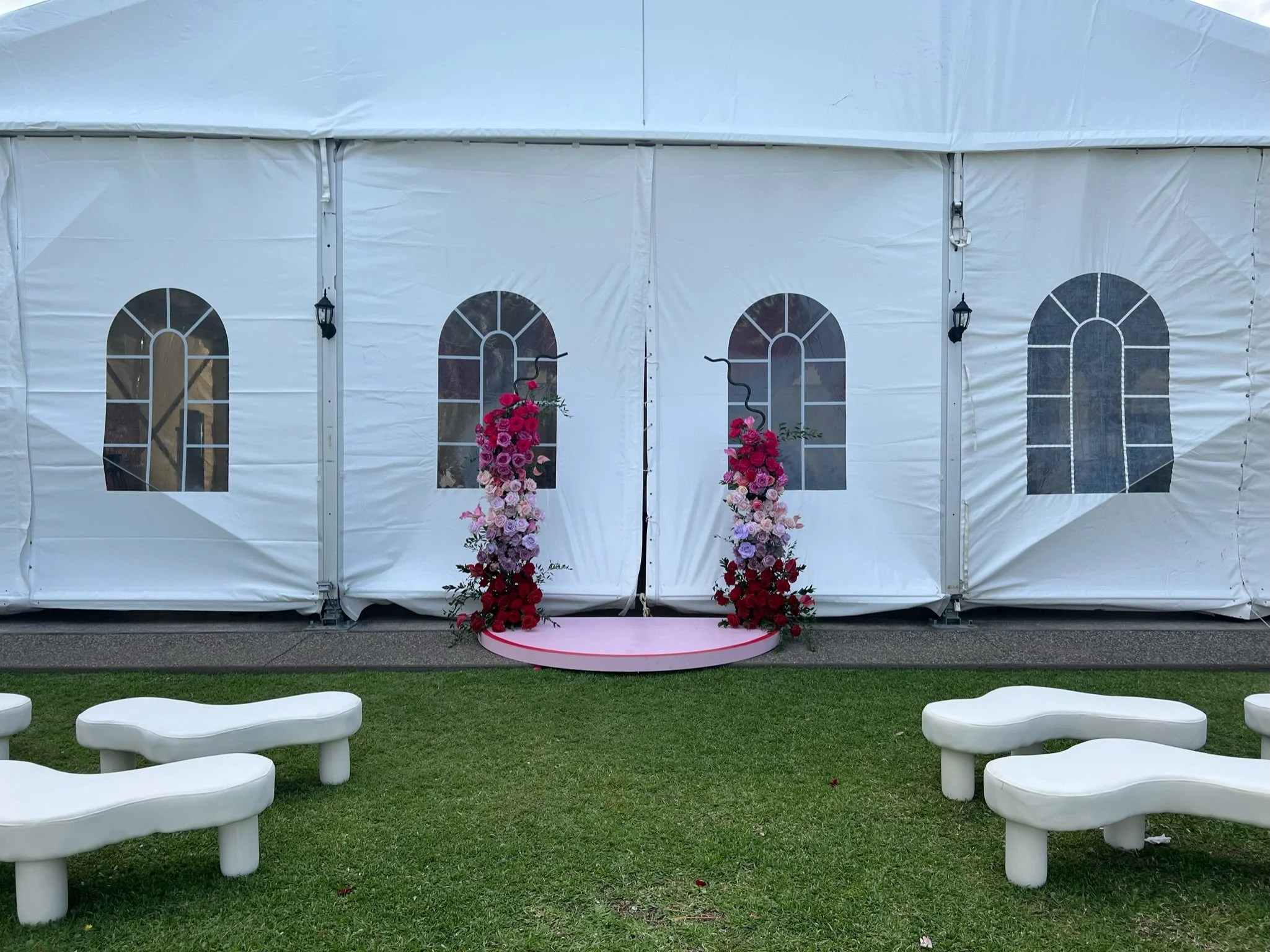 Wedding altar with pink and red flowers, draped in white, with white benches on green grass.