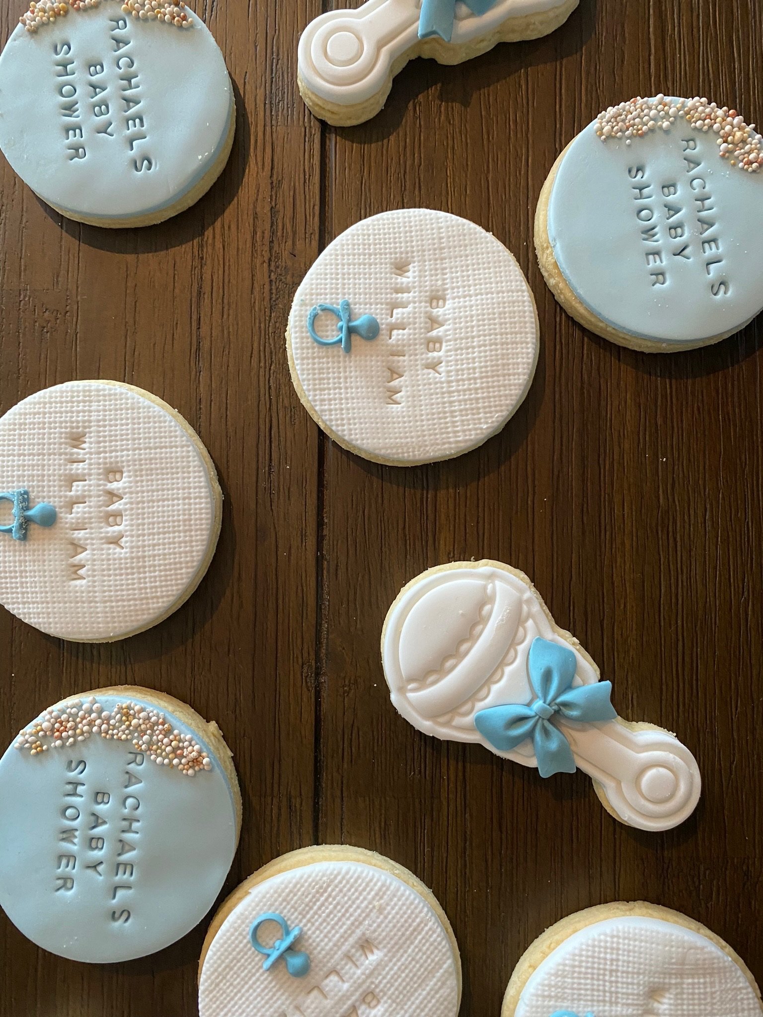 Baby shower cookies decorated with blue and white icing, some with the words 'Welcome Baby' and others with the name 'Rachel's Baby Shower,' some with baby-related designs like a rattle and a pacifier.