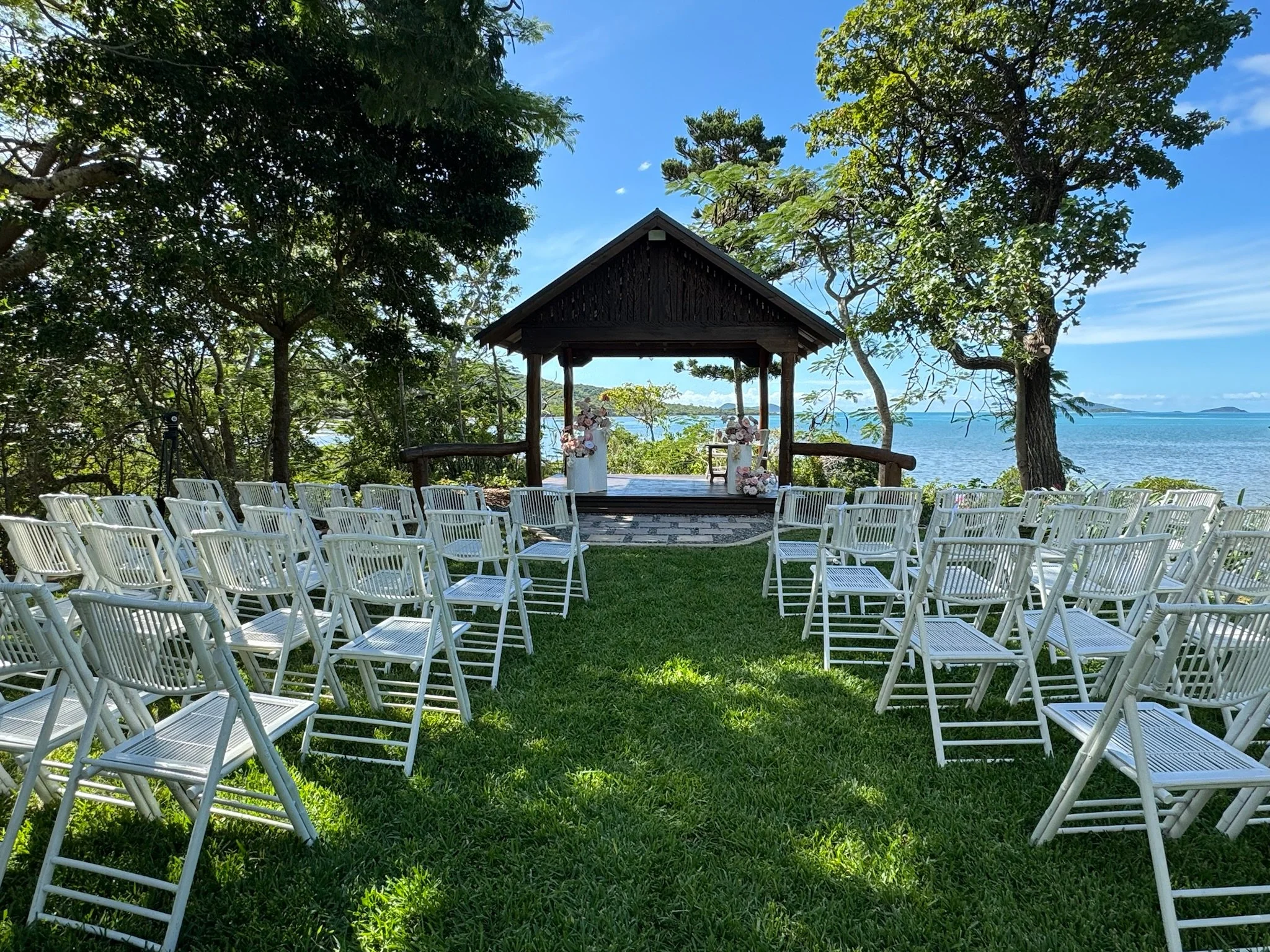 An outdoor wedding setup with white chairs arranged in rows, facing a small wooden gazebo decorated with flowers, overlooking a body of water with trees and islands in the background.