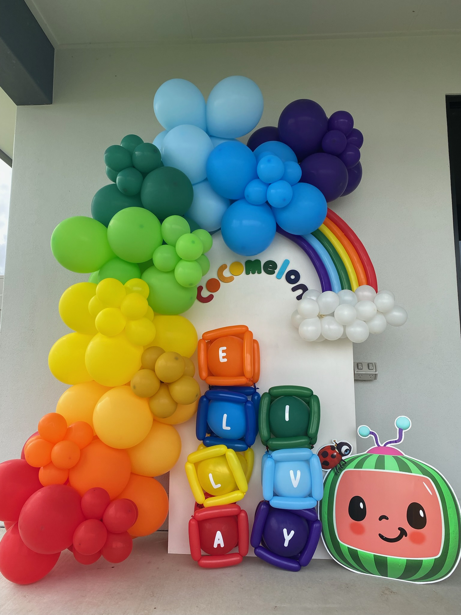 Colorful balloon decoration with a rainbow, the words "Welcome" and "ELOY" spelled out with balloons, and a cartoon watermelon character with a ladybug on its side.